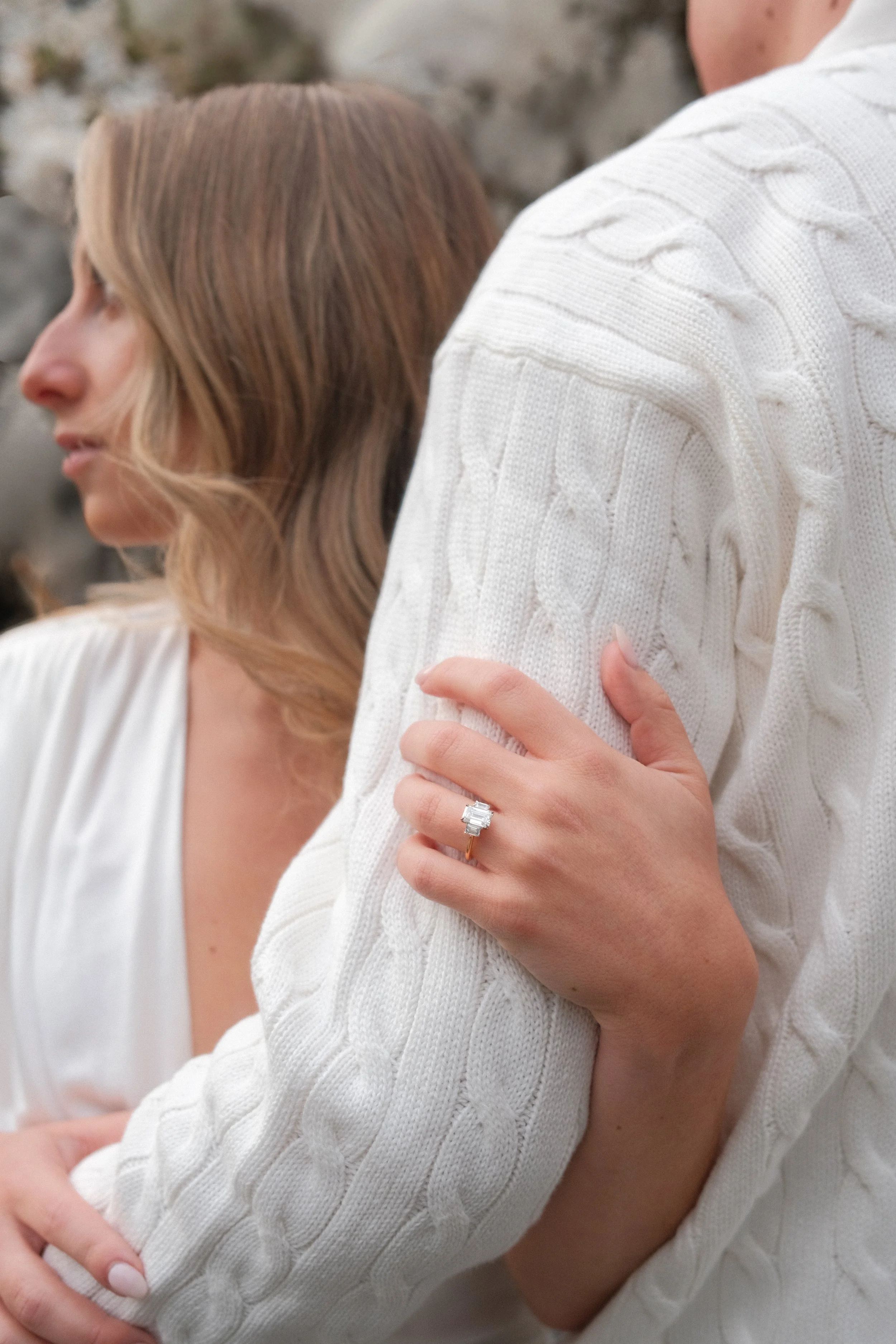 Close-up of a woman in a white cable knit sweater showing her left hand with a square-cut diamond engagement ring on her finger, with her arm resting on another person wearing a similar sweater.