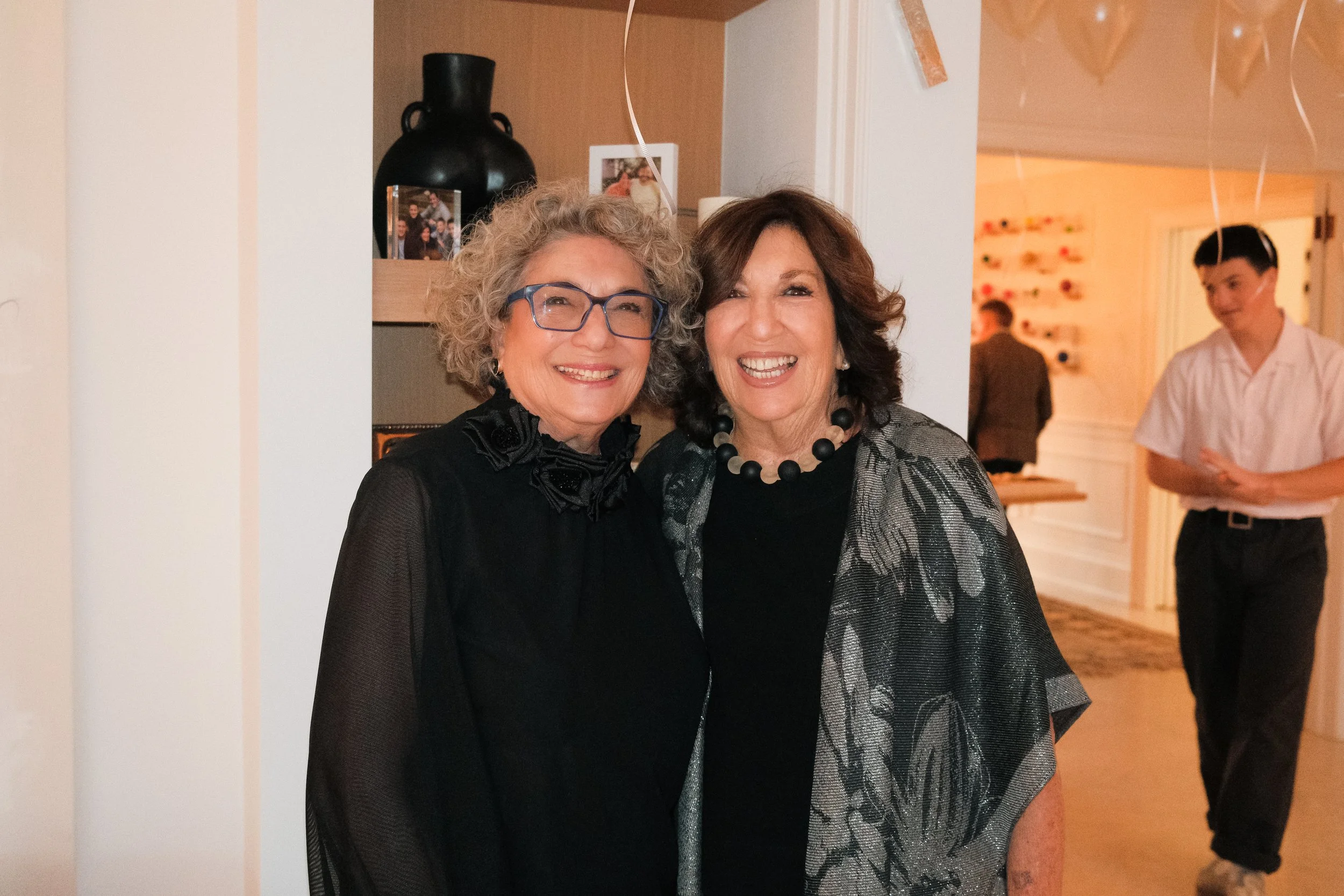 Two women, smiling, standing indoors at a social gathering. One has curly gray hair and glasses, and the other has short, dark, wavy hair and a large bead necklace.