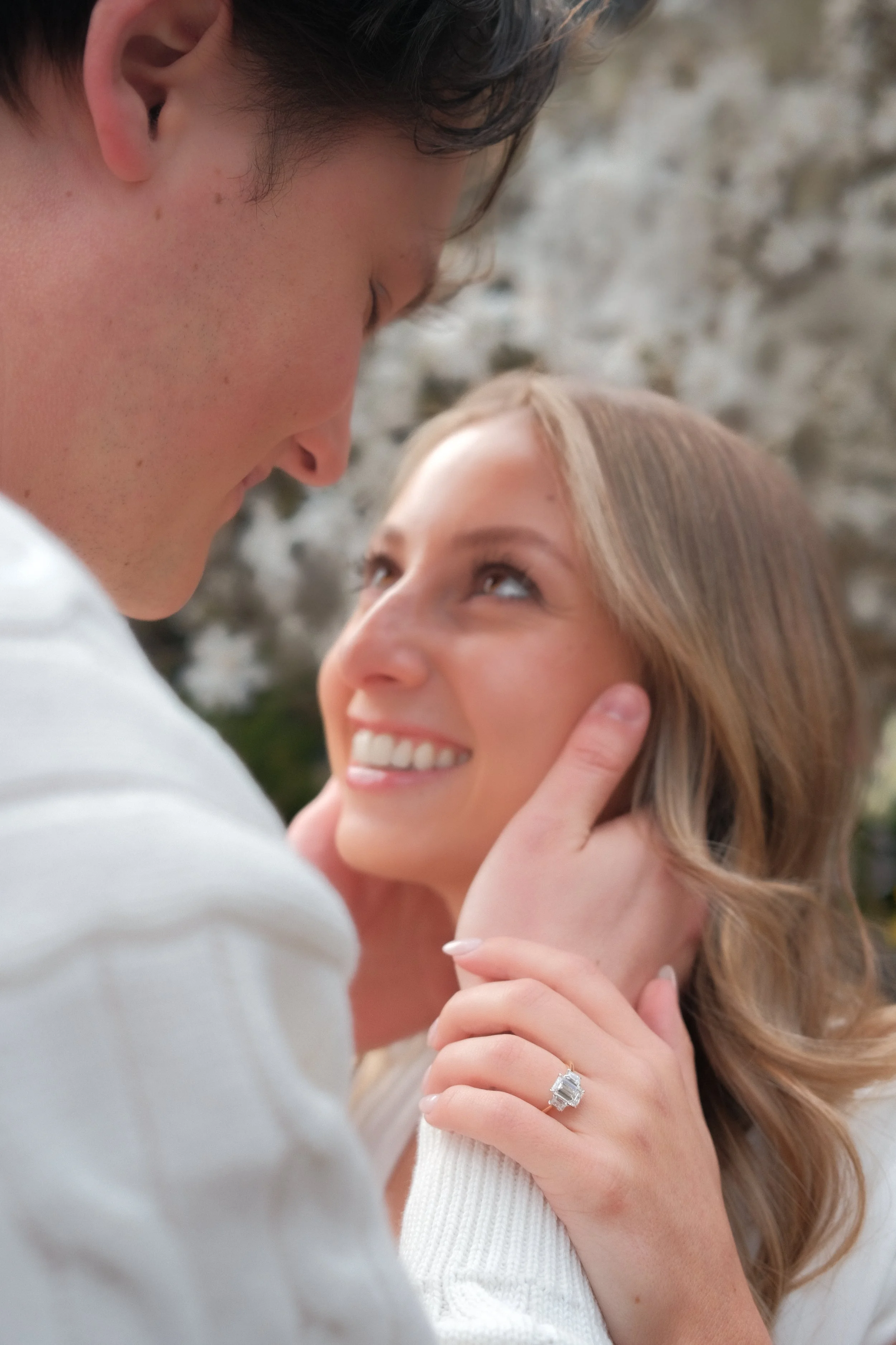 A couple shares an intimate moment outside, with the man gently holding the woman's face and both smiling warmly at each other.