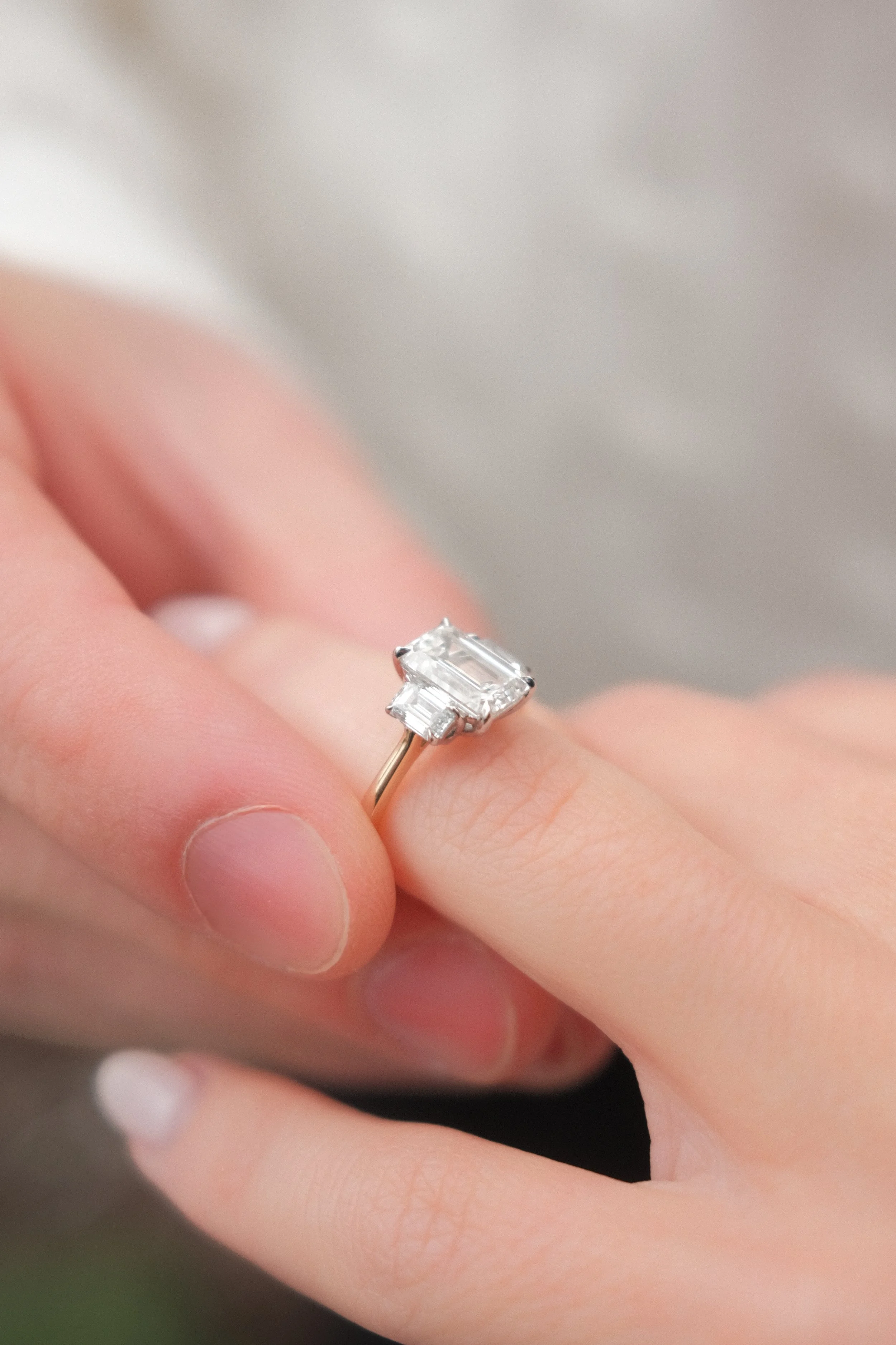 Close-up of a hand with a diamond engagement ring featuring a large rectangular center stone and baguette side stones on a gold band.