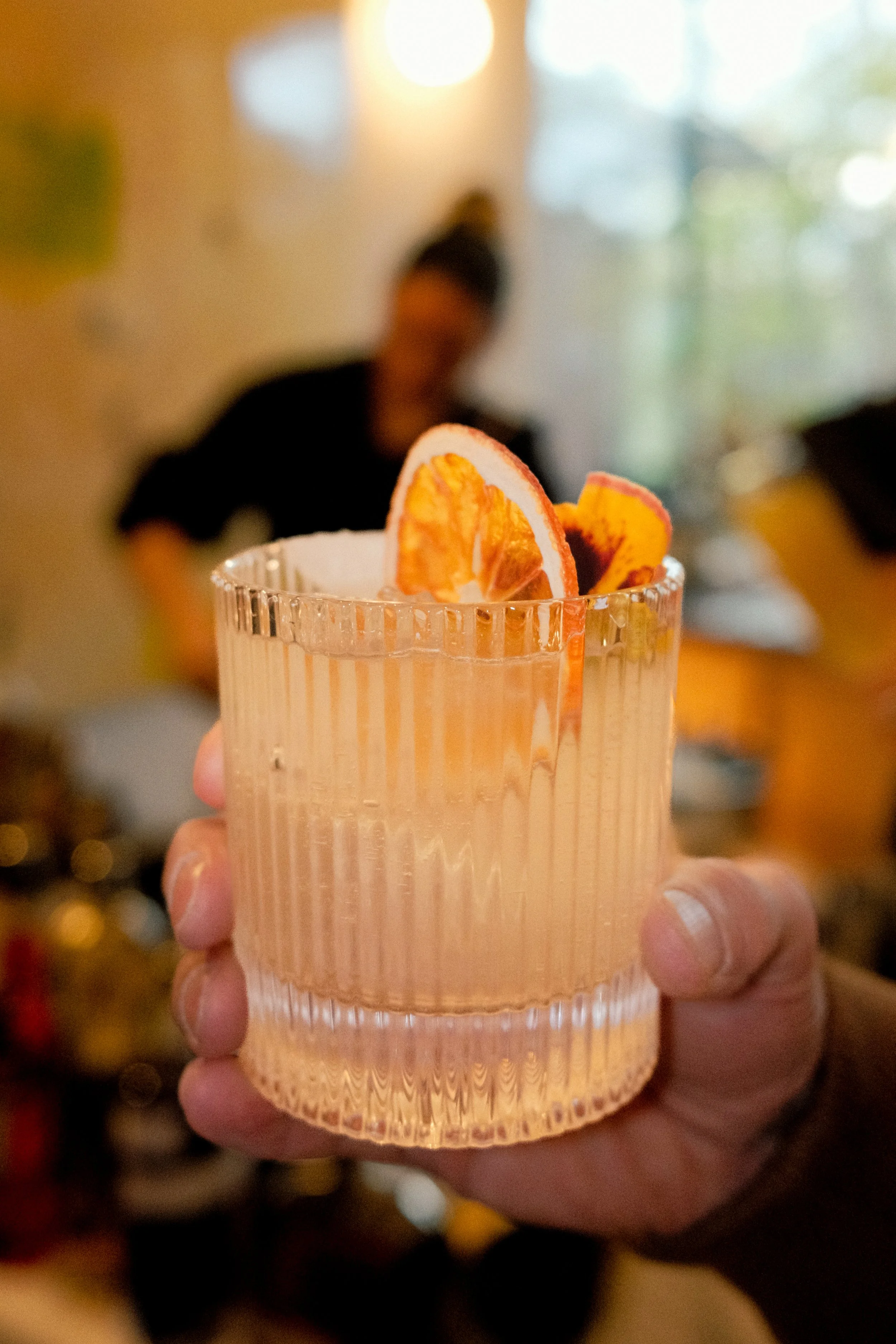 A person holding a glass with dried citrus slices garnish. In the background, a blurred woman is standing near a window.