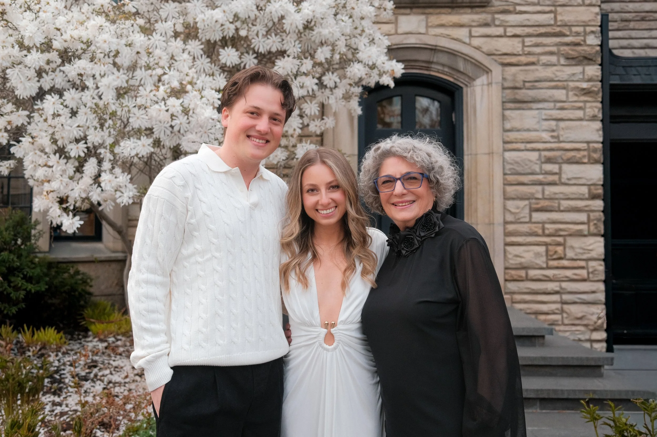 Three women smiling in front of a brick house with blooming white flowers.