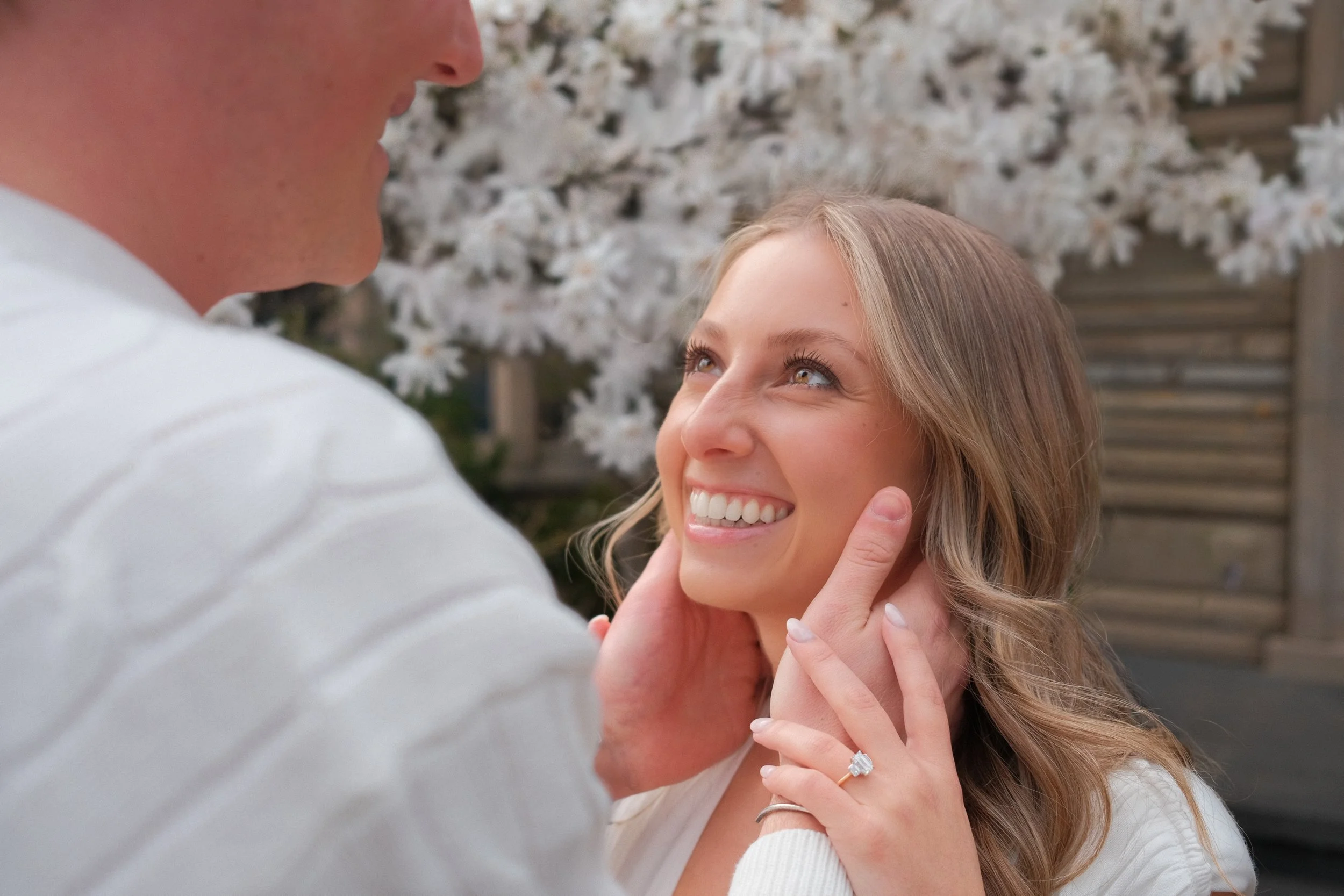 A woman and a man smiling at each other outdoors, with a background of white flowering trees. She is wearing a ring, and the man is gently holding her face.