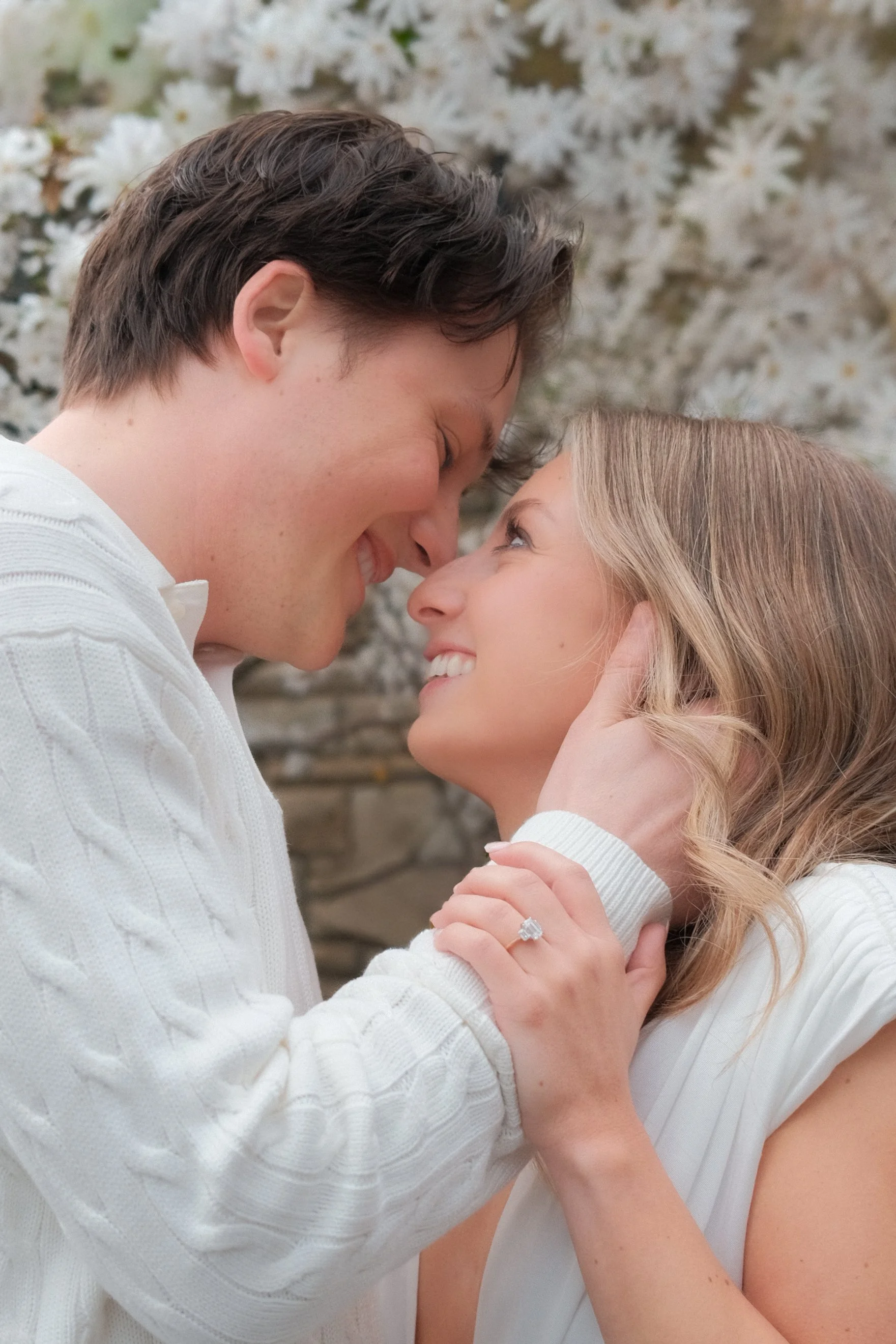 A couple with light skin faces close close, smiling and about to kiss in front of a flowers background