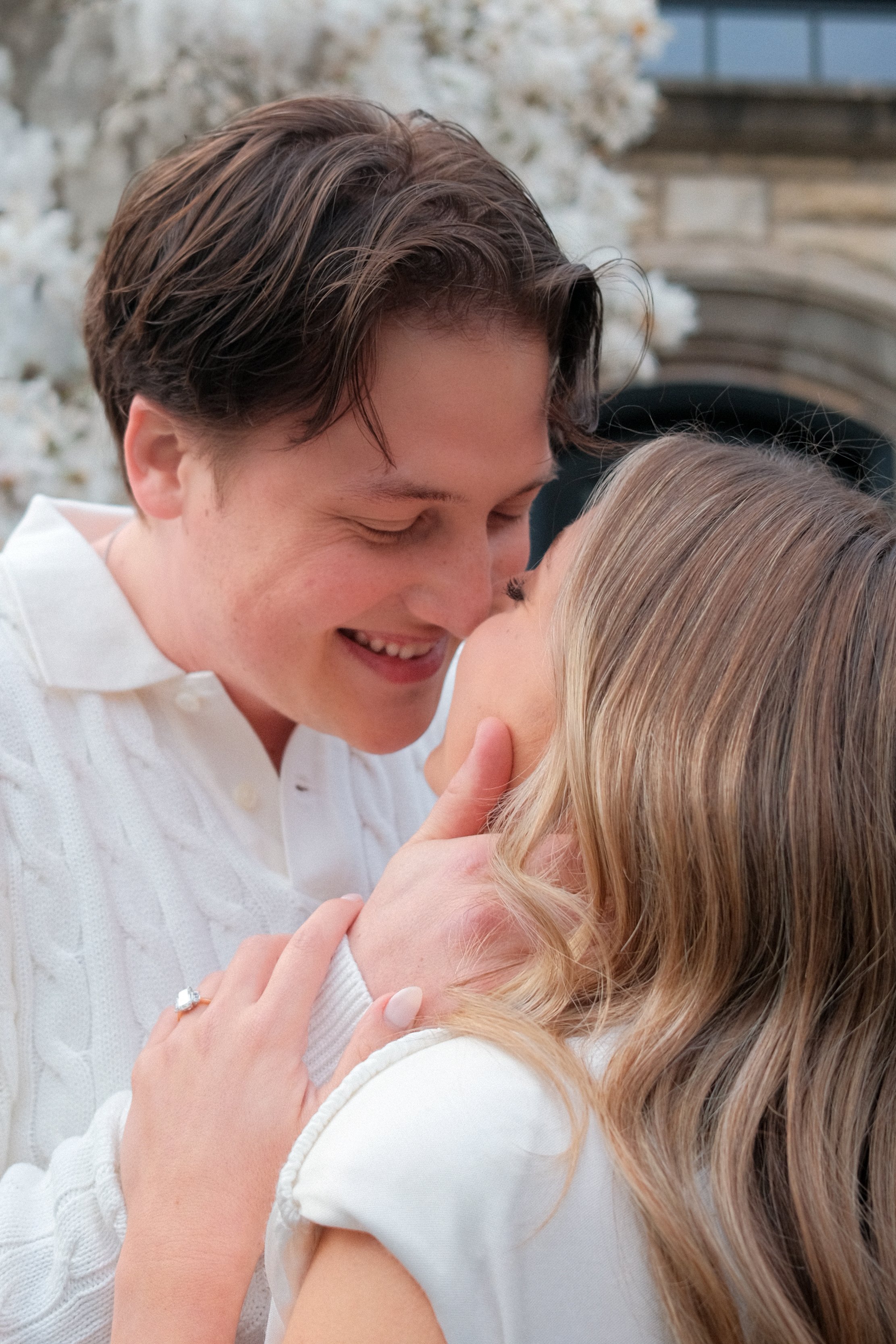 A young couple sharing a close, intimate moment outside, with the man smiling and the woman touching his face, surrounded by a blurred natural background.