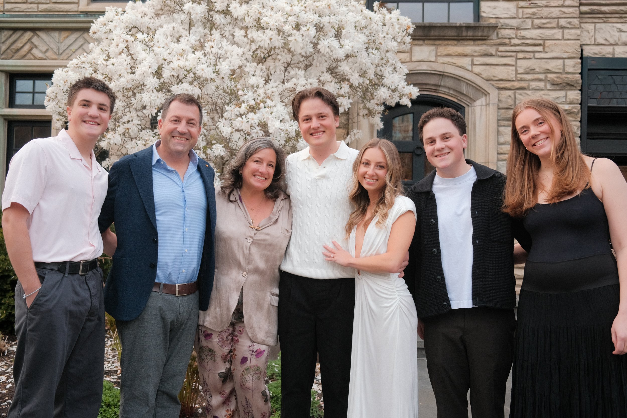 Group of seven people smiling in front of a blooming white tree with a house in the background.