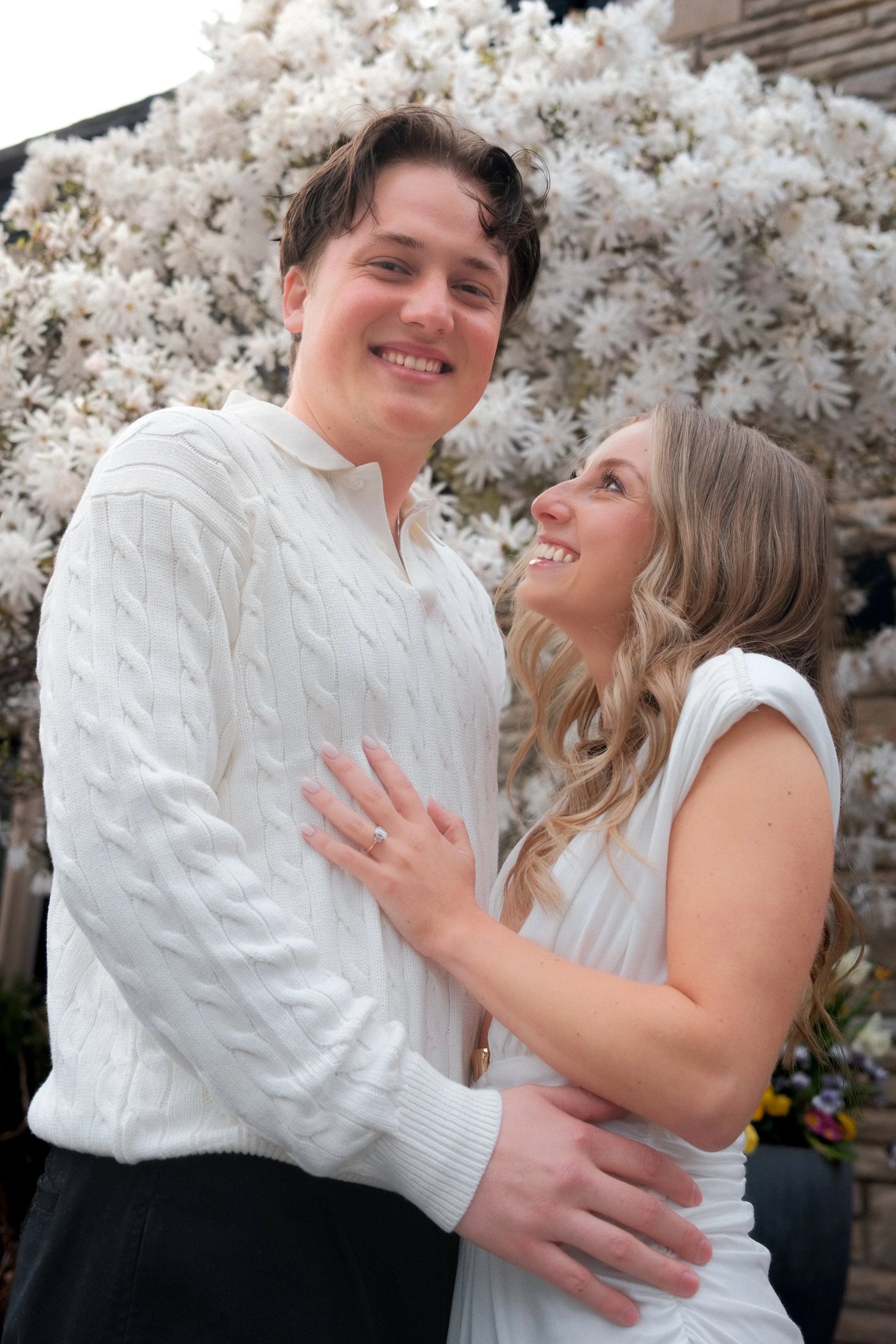 A young couple indoors, standing close together, smiling at each other with a white flowering plant in the background.
