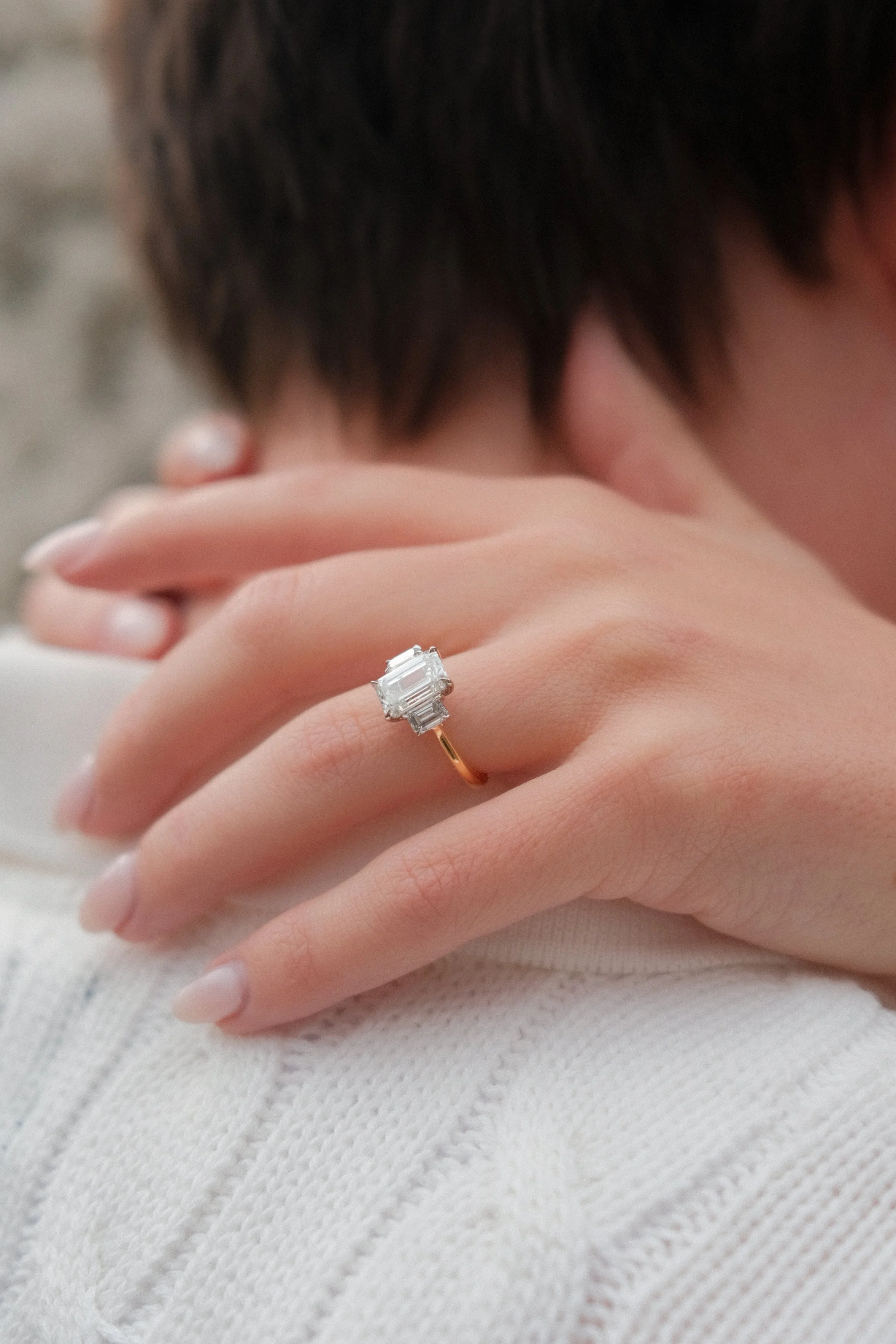 A close-up of a person's hand wearing a large rectangular diamond ring, resting on their chest with a blurred background.