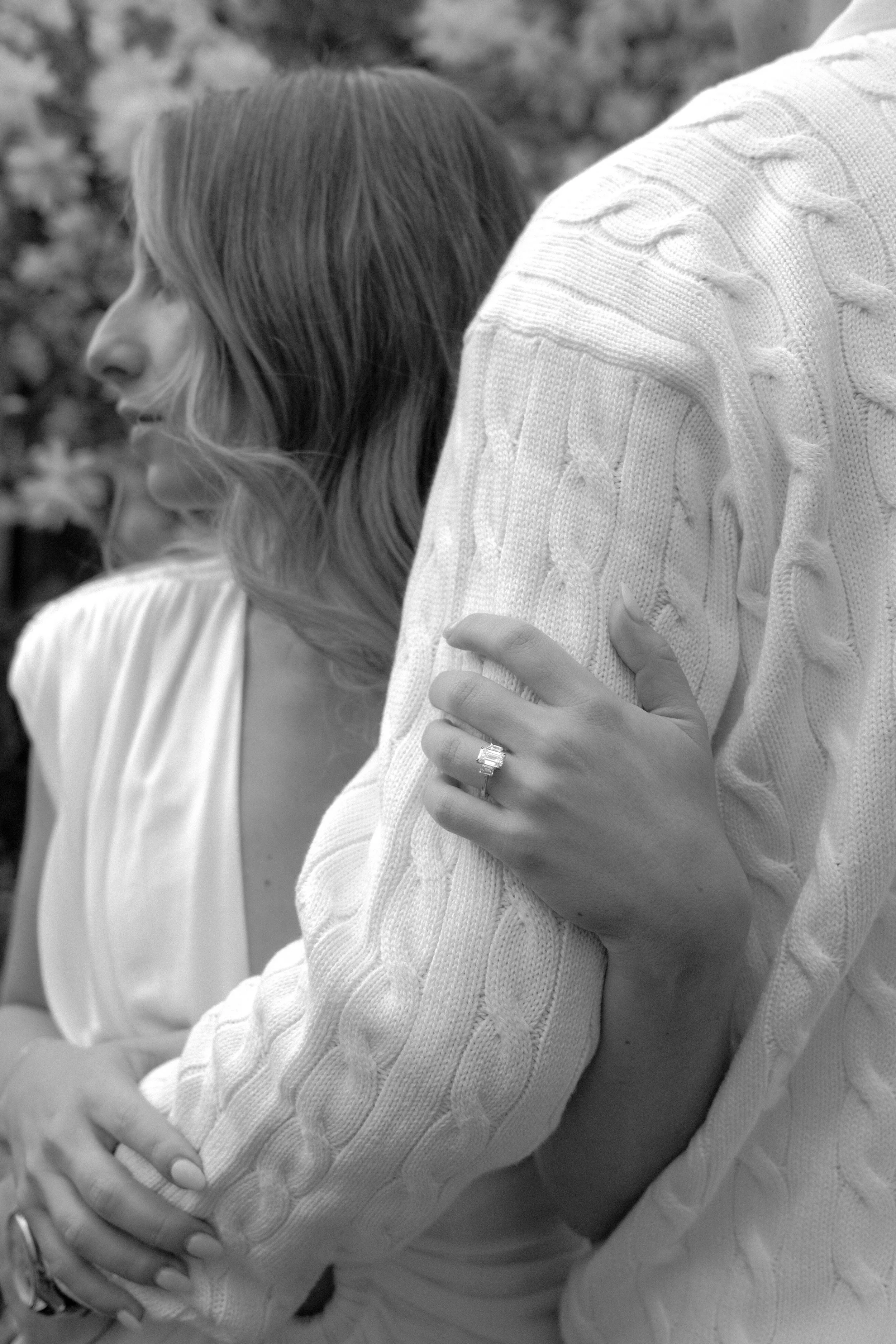 Close-up of two people, one woman looking at the left and another woman showing her arm, wearing a cable-knit sweater, with rings on fingers, against a blurred outdoor background.