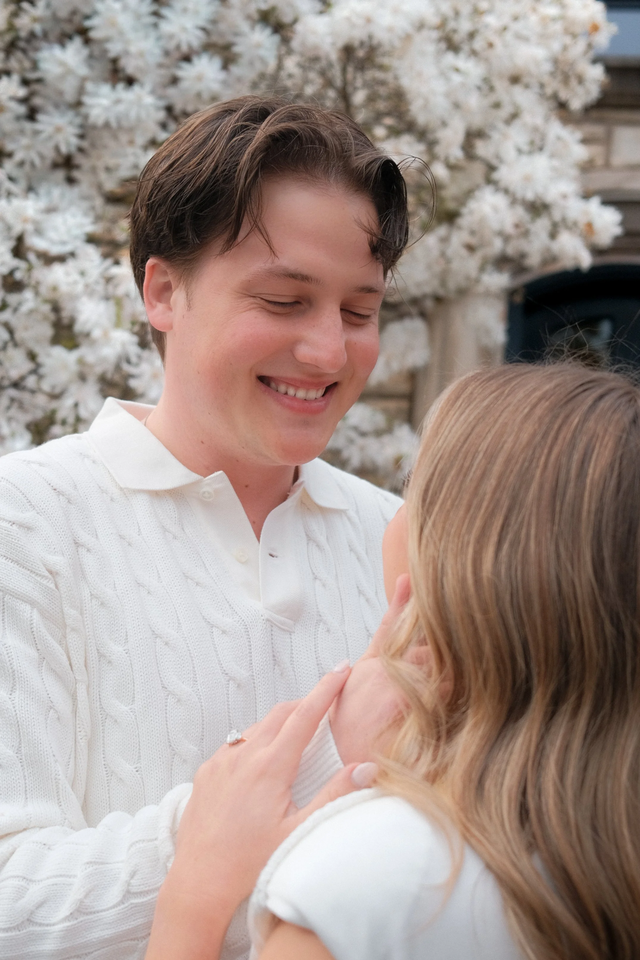 A young man with dark hair smiling with his eyes closed, wearing a white sweater, holding a small girl with long light brown hair, outdoors with white flowering trees and a blue sky in the background.
