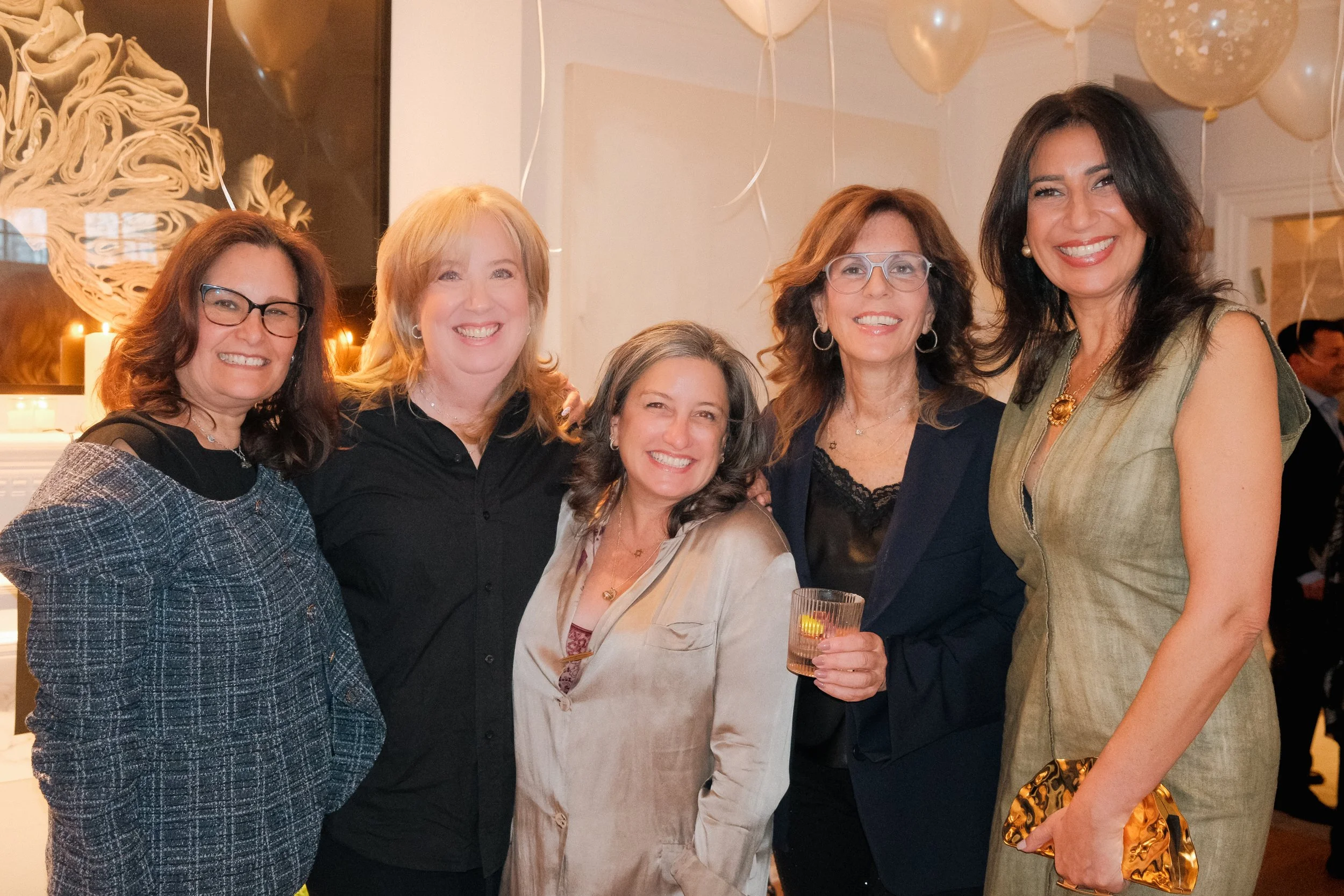 A group of six women smiling indoors at a celebration, with balloons and decorations in the background.