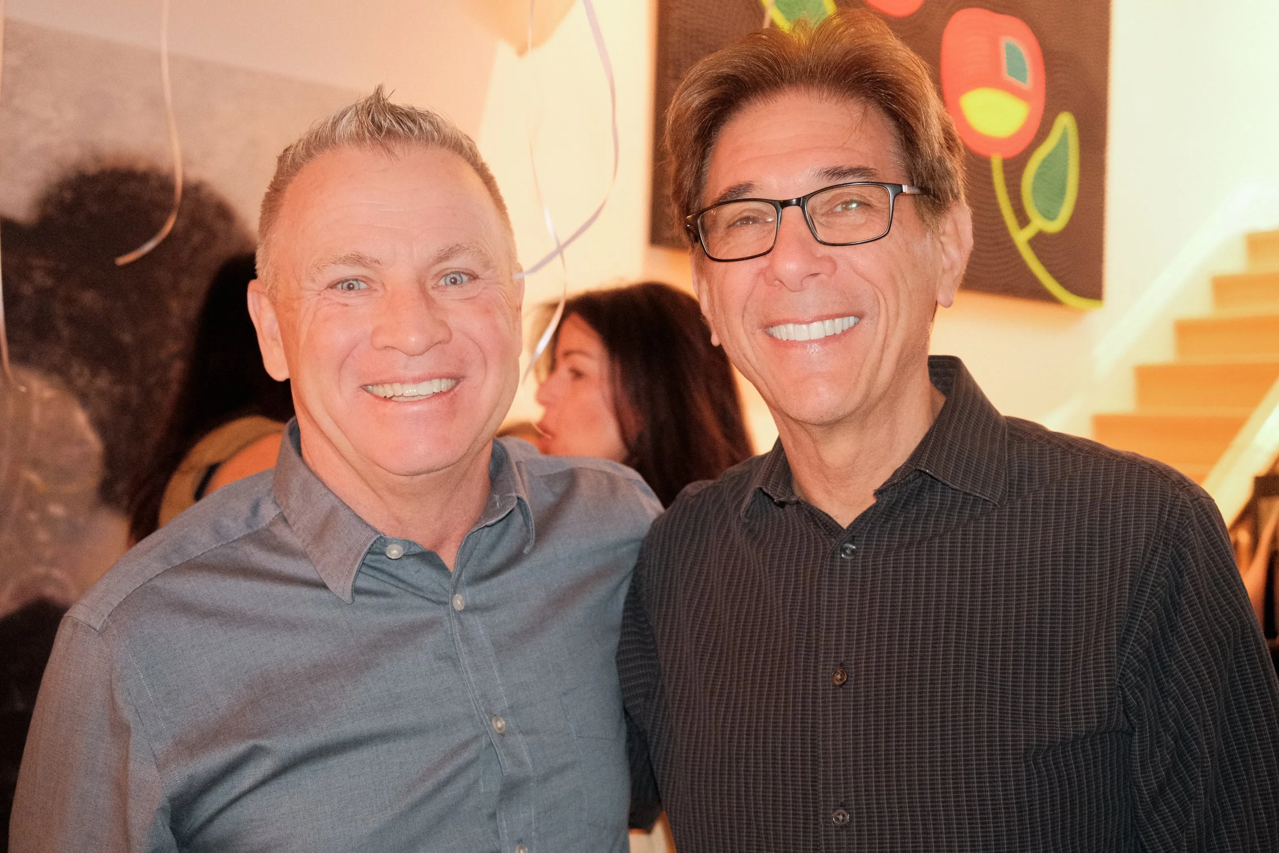 Two middle-aged men smiling and standing close together at an indoor event, with other people in the background and colorful artwork on the wall.