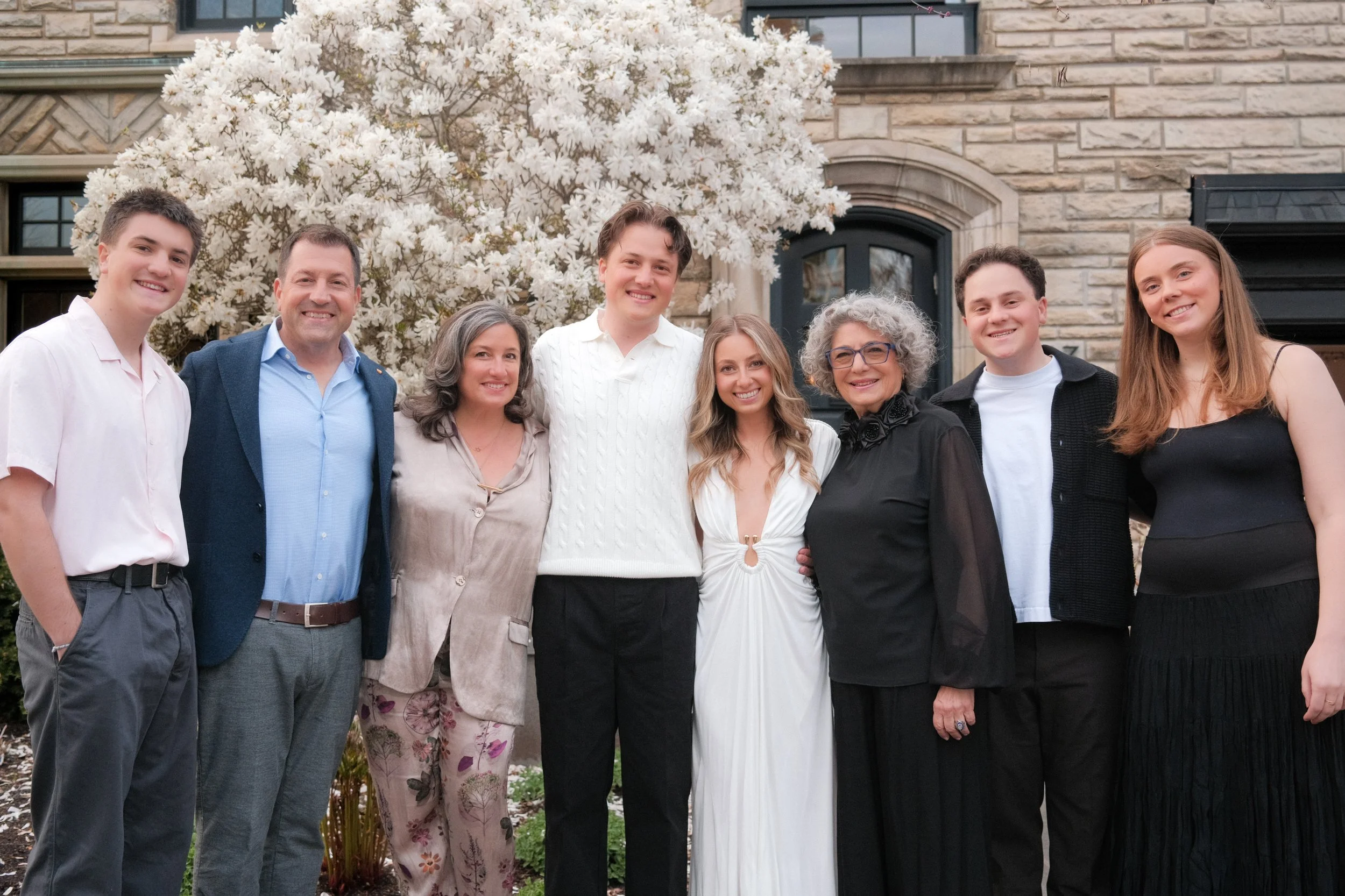 A group of nine people standing outside in front of a flowering tree and a house, smiling at the camera.