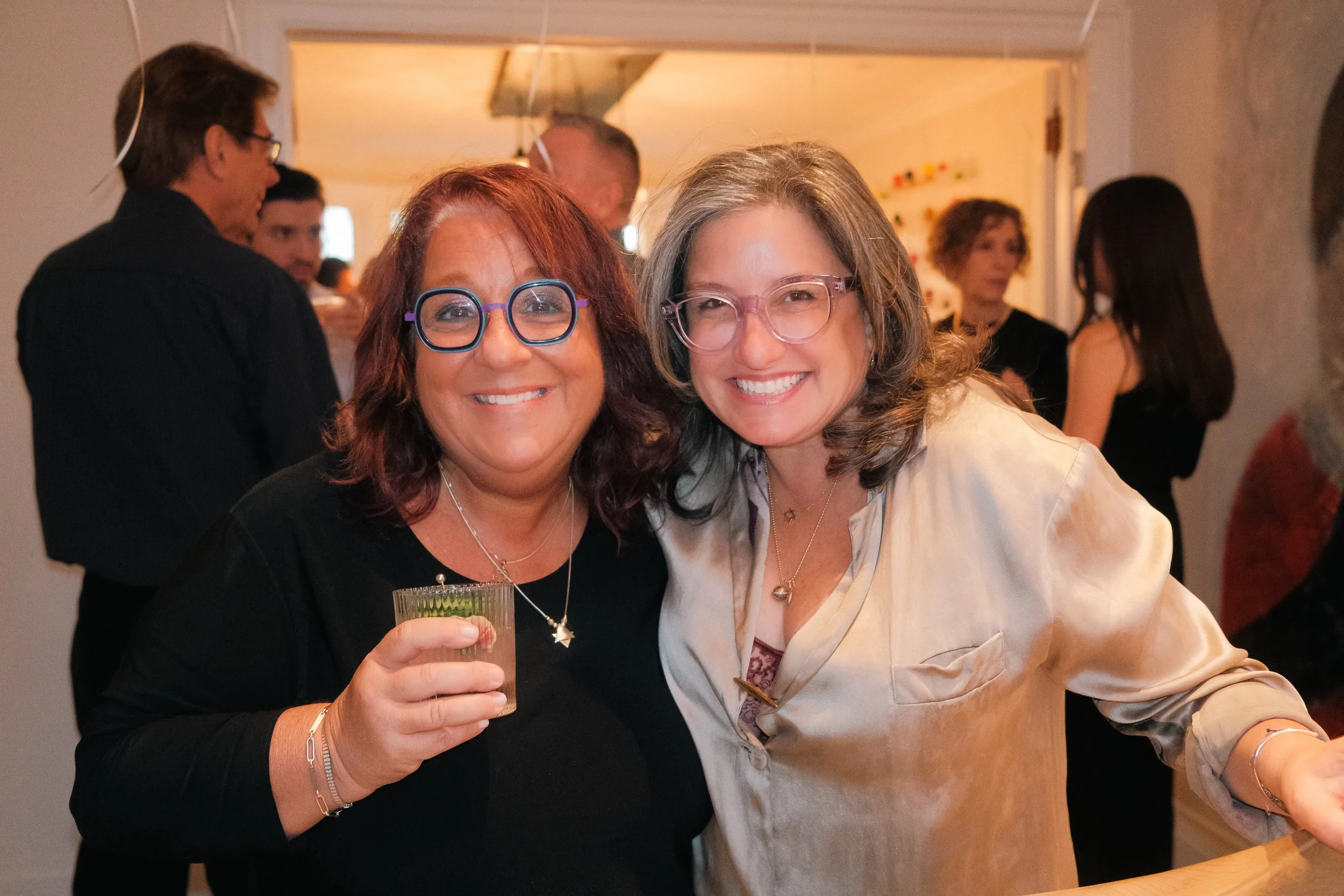 Two women smiling at a social gathering, one holding a drink, with other people blurred in the background.