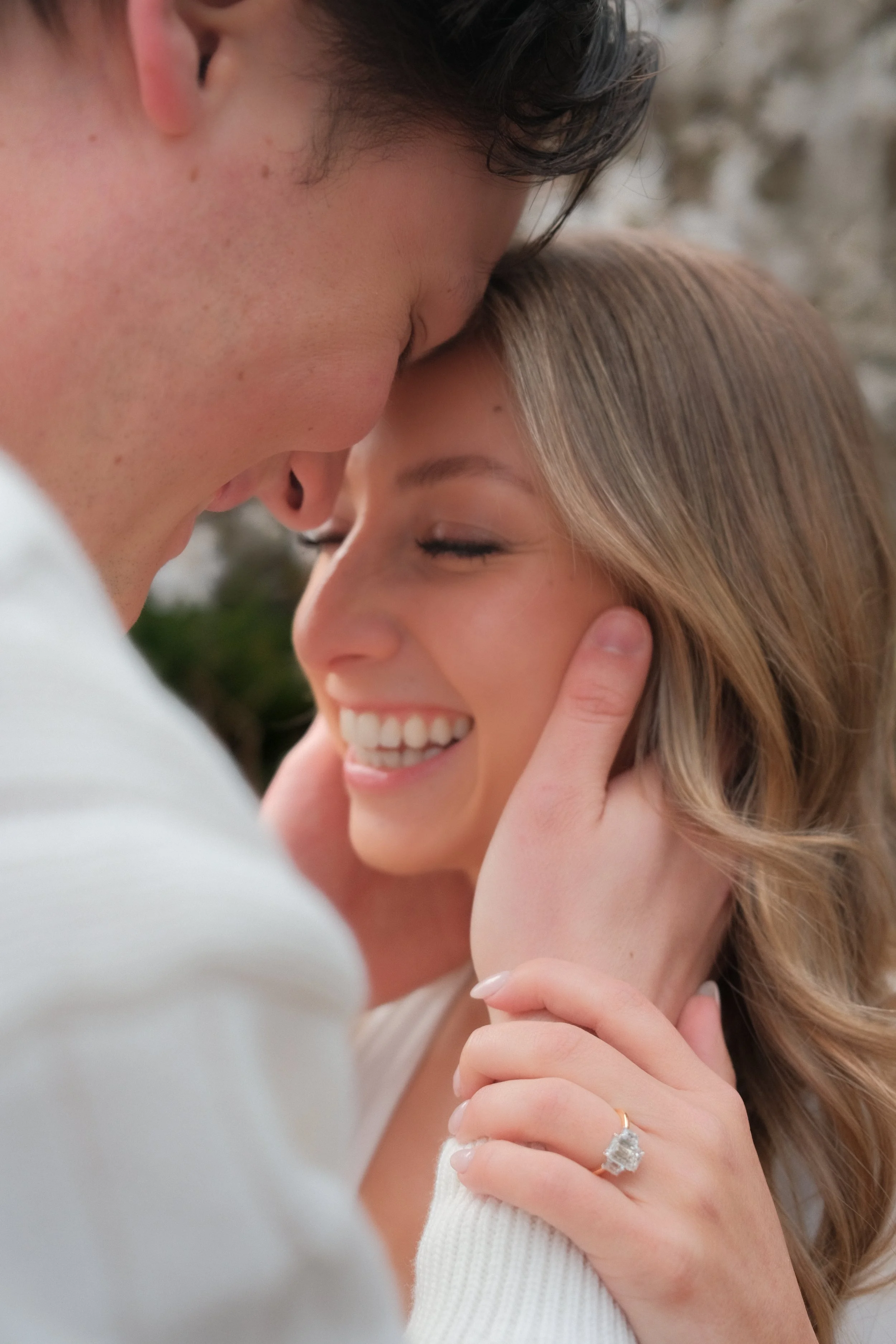 Close-up of a couple smiling with heads touching, the man gently holding the woman's face, wearing an engagement ring, outdoors.