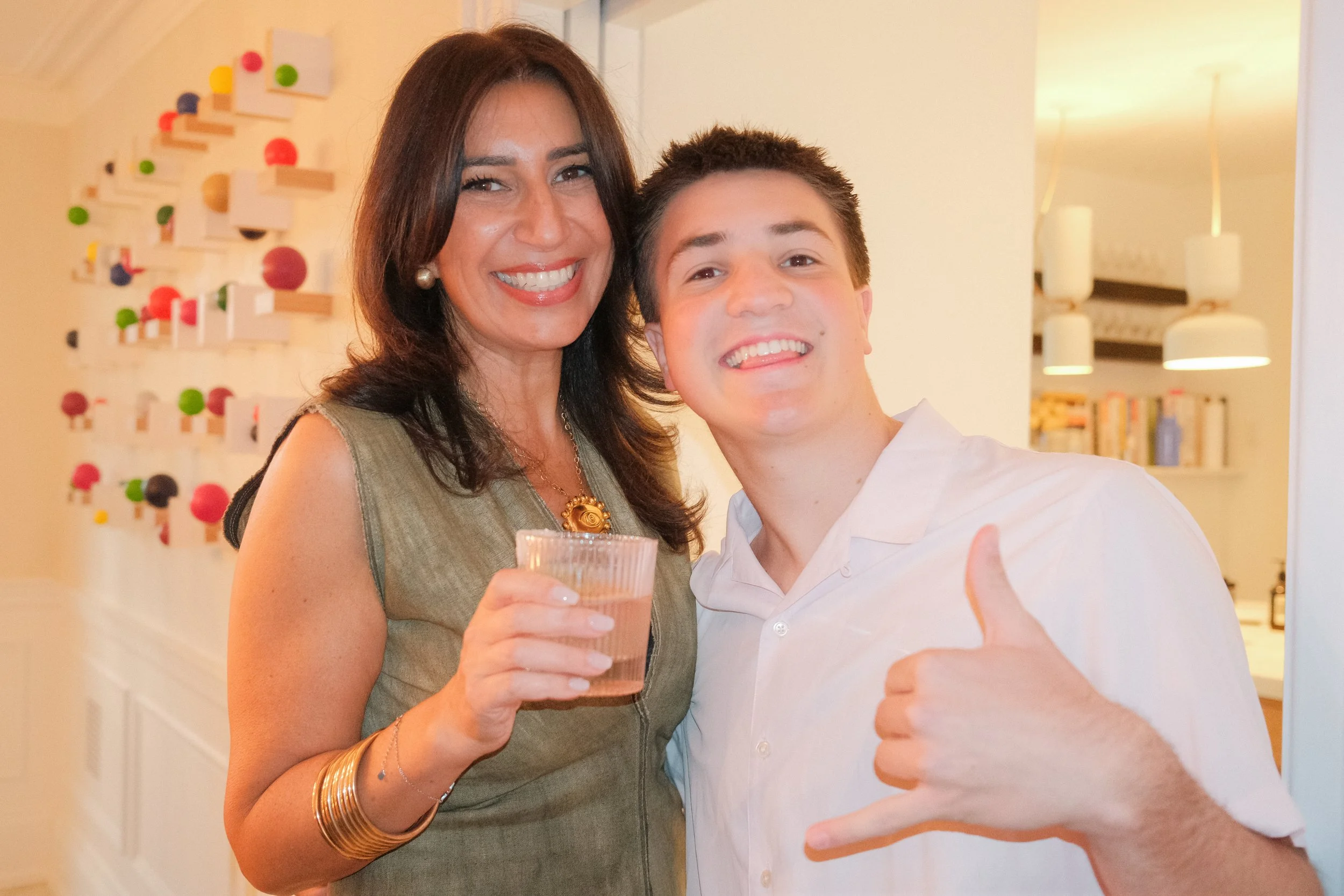 A woman and a boy are smiling and posing for a photo at a social gathering. The woman has dark brown hair and is wearing a green dress with a gold necklace and earrings, and is holding a glass of drink. The boy has short dark hair and is wearing a wh