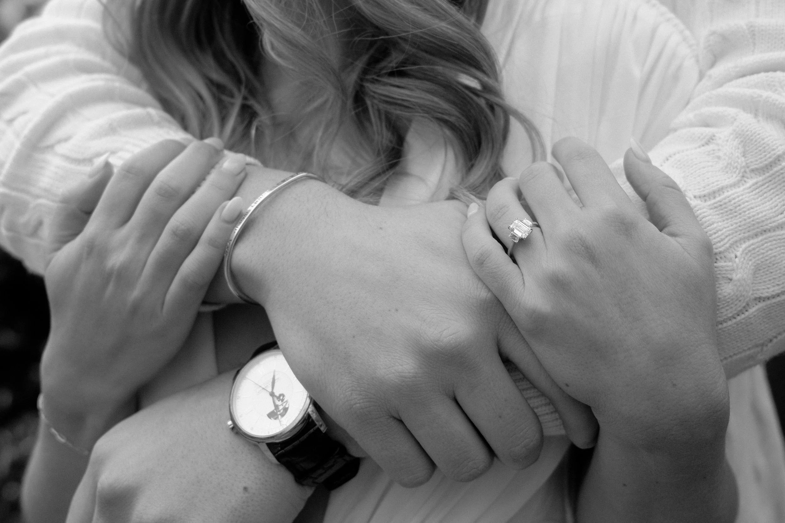 A woman with curly hair resting her head on her crossed arms on a surface, wearing a watch and rings on her fingers.