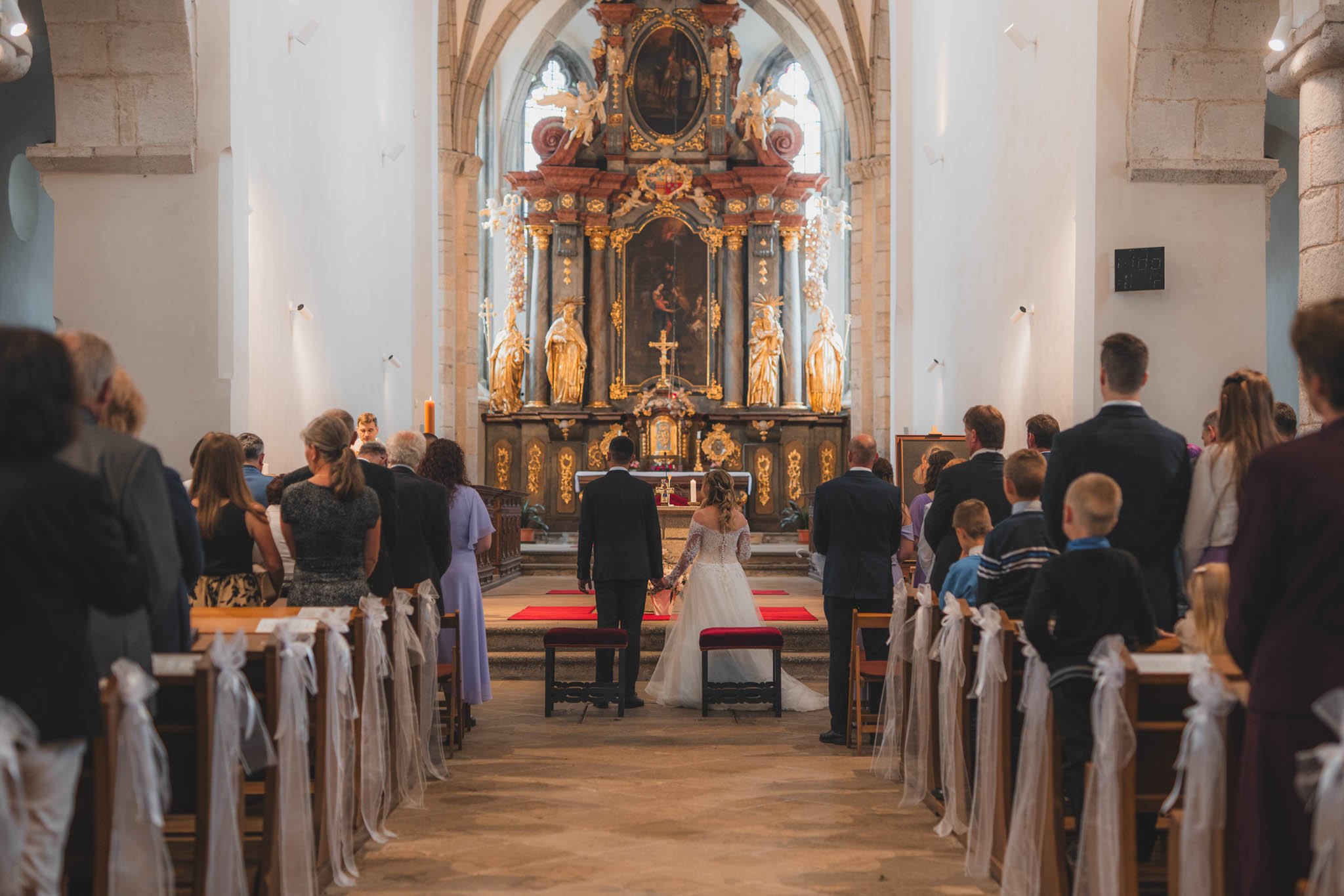 Nevěsta a ženich stojí u oltáře v kostele, obklopeni svatebními hosty.