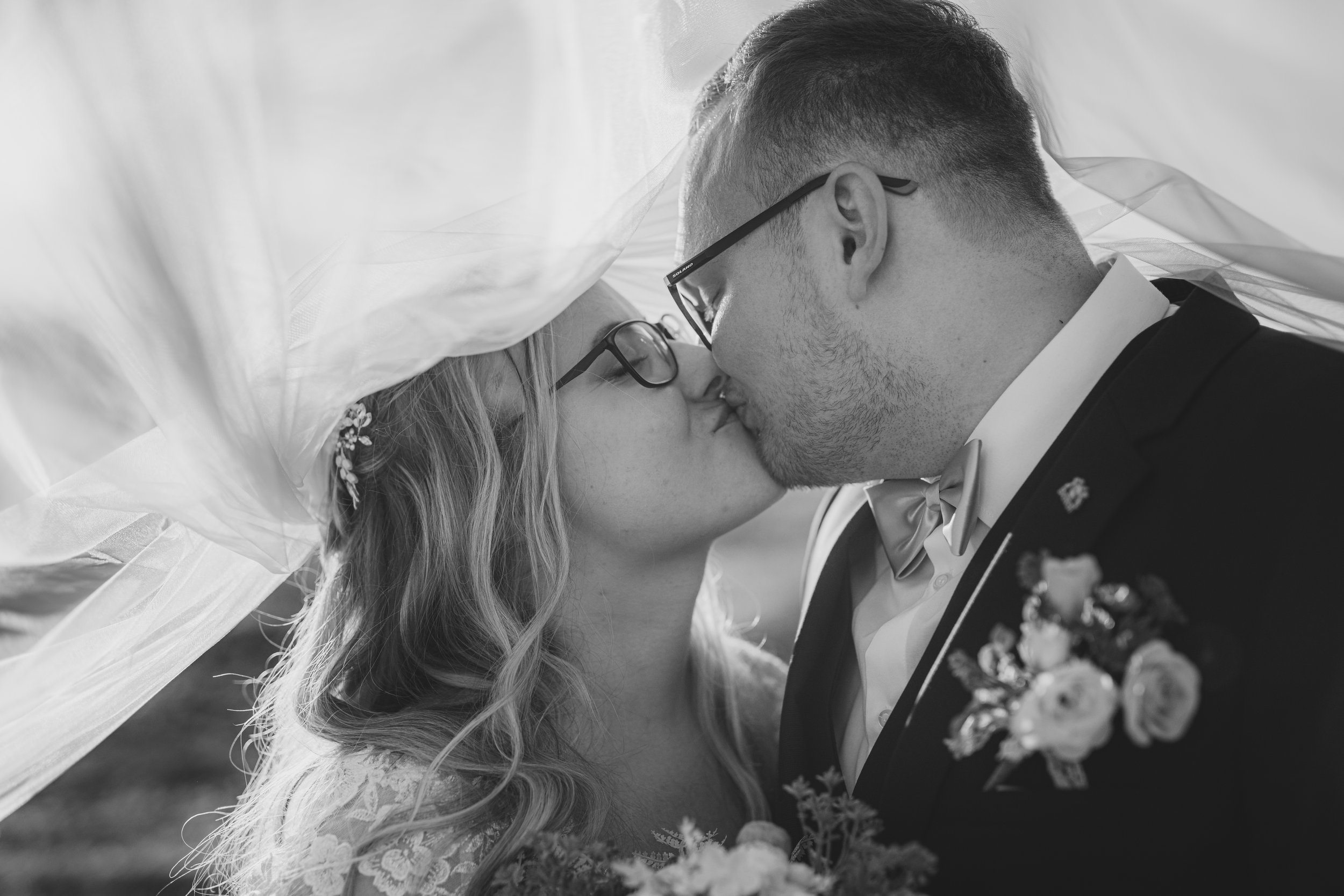 Černobílá fotografie nevěsty a ženicha, jak se líbají pod závojem nevěsty během svatby.