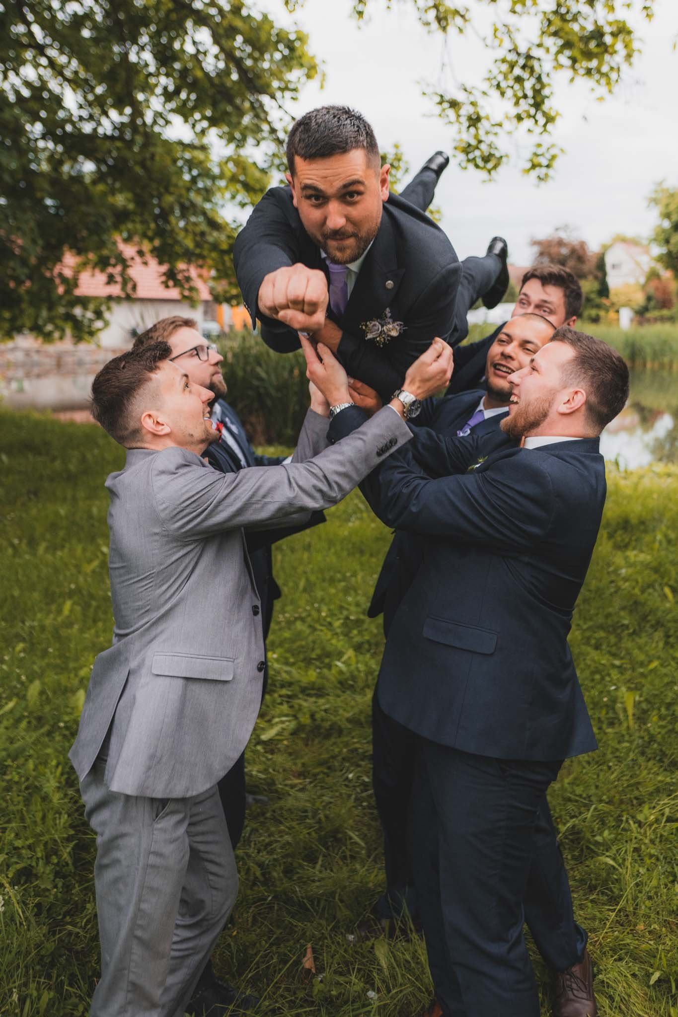 Skupina pěti mužů v oblecích venku zvedá muže v obleku do vzduchu, kolem jsou stromy a tráva.