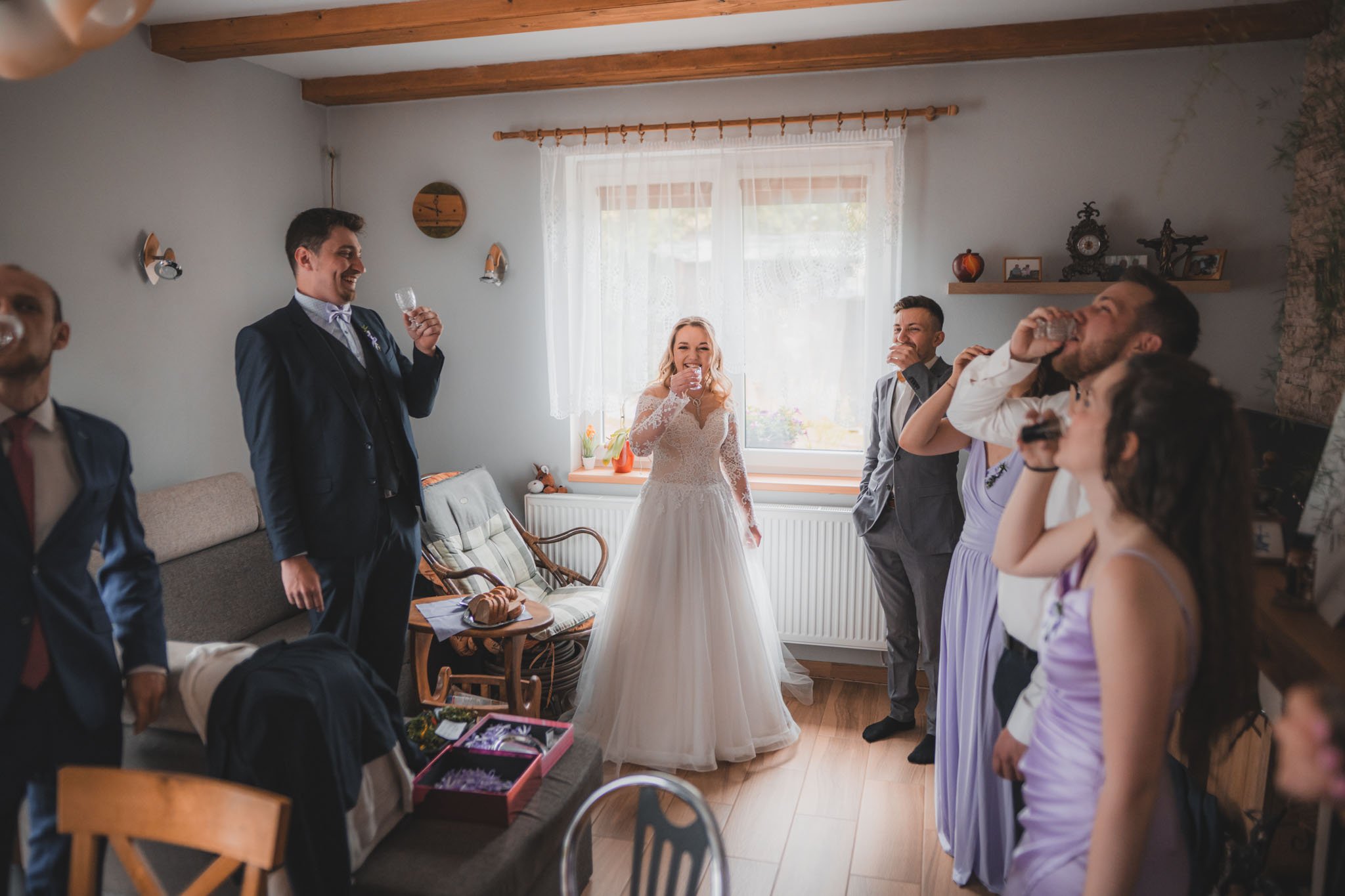 Skupina lidí slaví uvnitř místnosti s nevěstou v bílých svatebních šatech, obklopená přáteli a rodinou, kteří pijí a usmívají se.