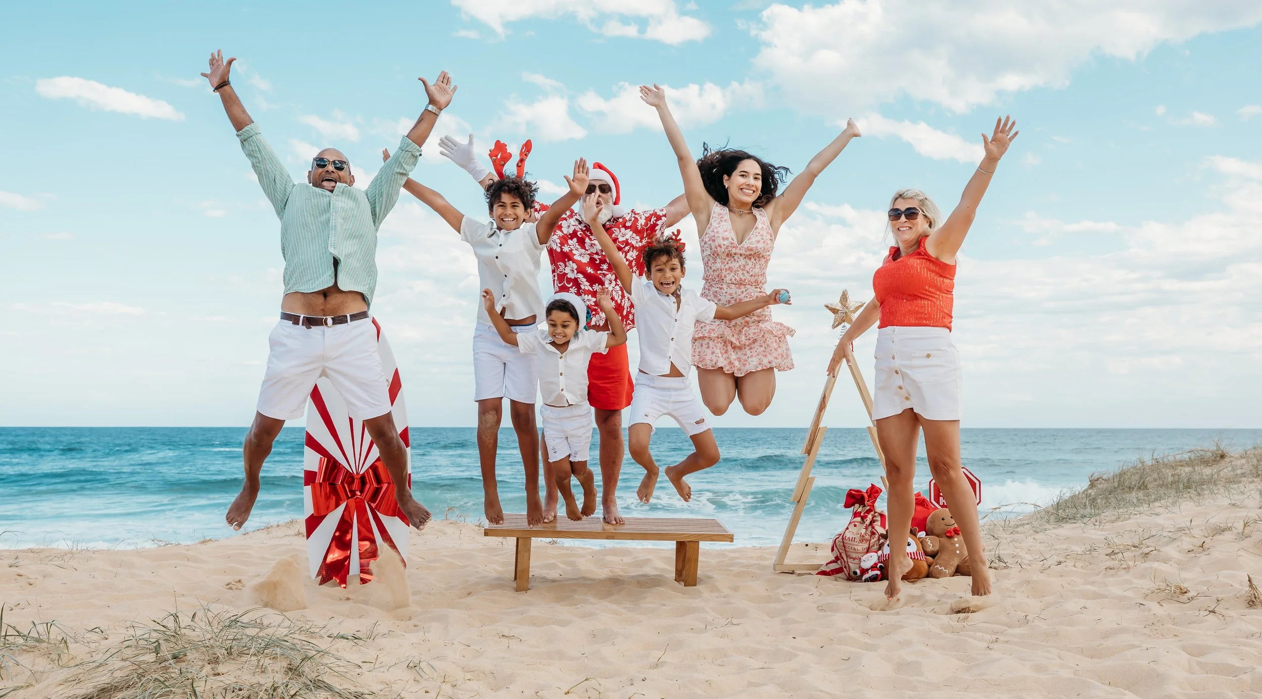 A multigenerational family celebrating Christmas at the beach, with people jumping and smiling under a blue sky.