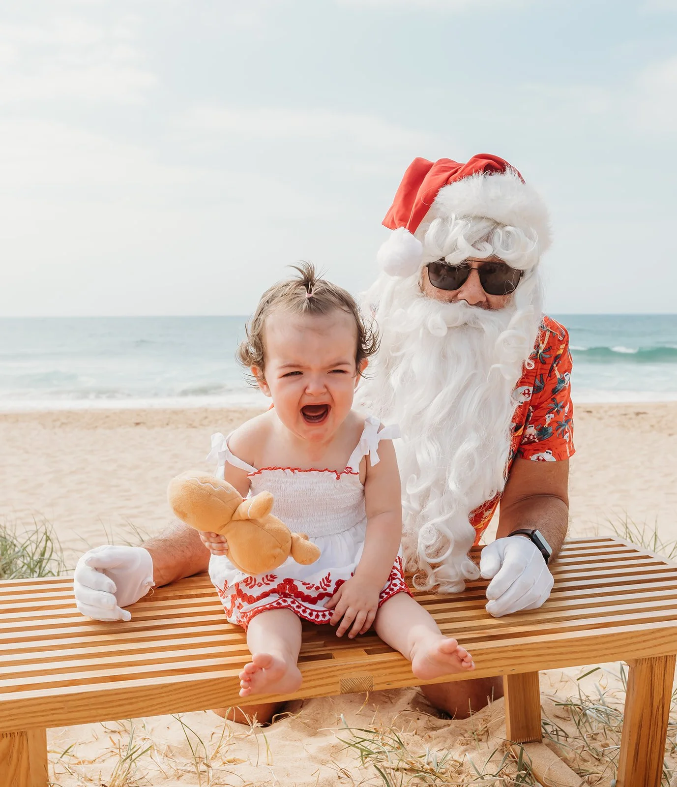 A man dressed as Santa Claus with sunglasses and a festive shirt sitting on a wooden bench at the beach, holding a young girl in a white dress with red embroidery, who is holding a teddy bear, both sitting on a wooden bench with sand and grass beneath them, with the ocean and sky in the background.