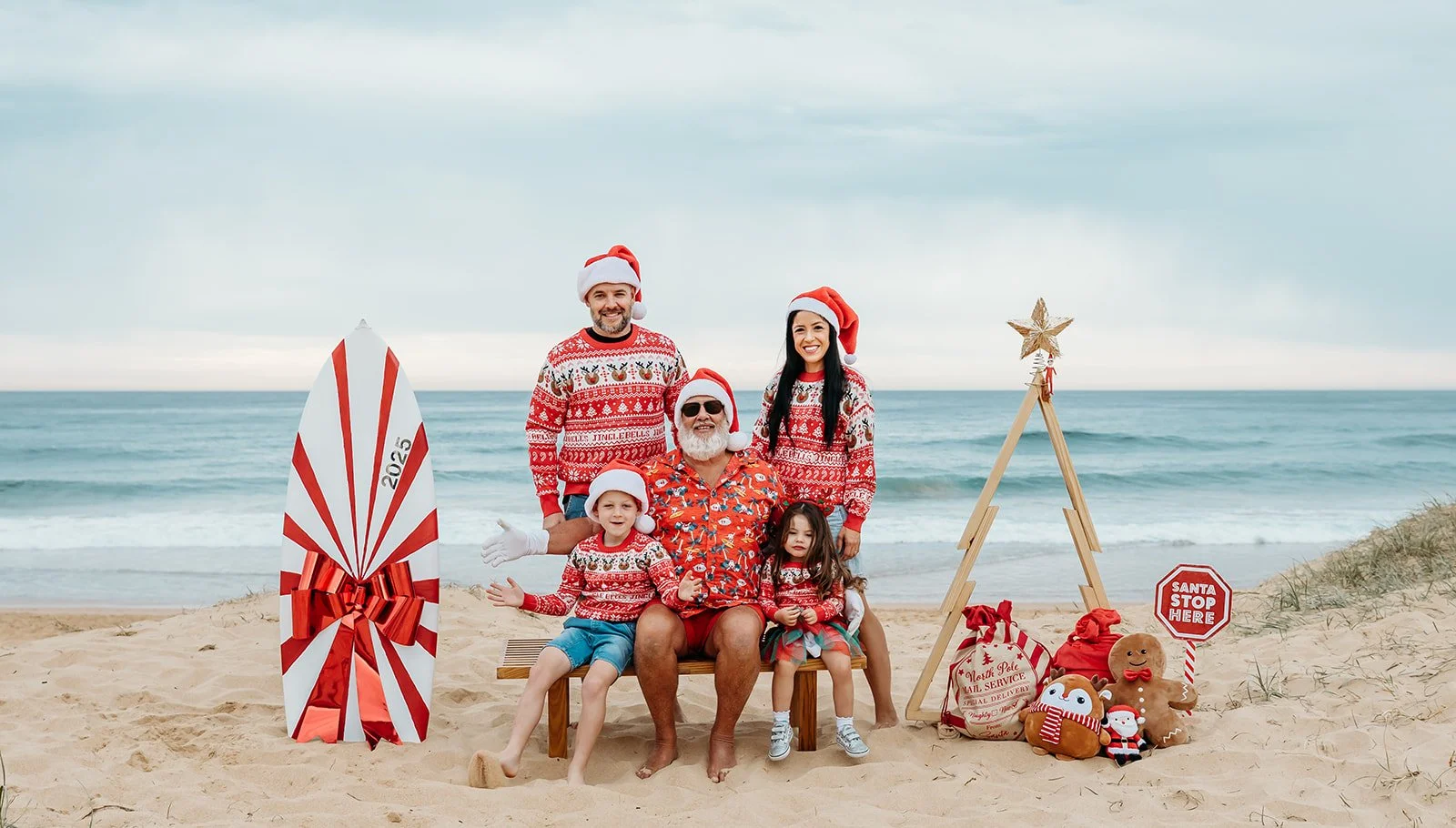 Family dressed in Christmas sweaters and Santa hats sitting and standing on a beach with holiday decorations, surfboard, and ocean in the background.