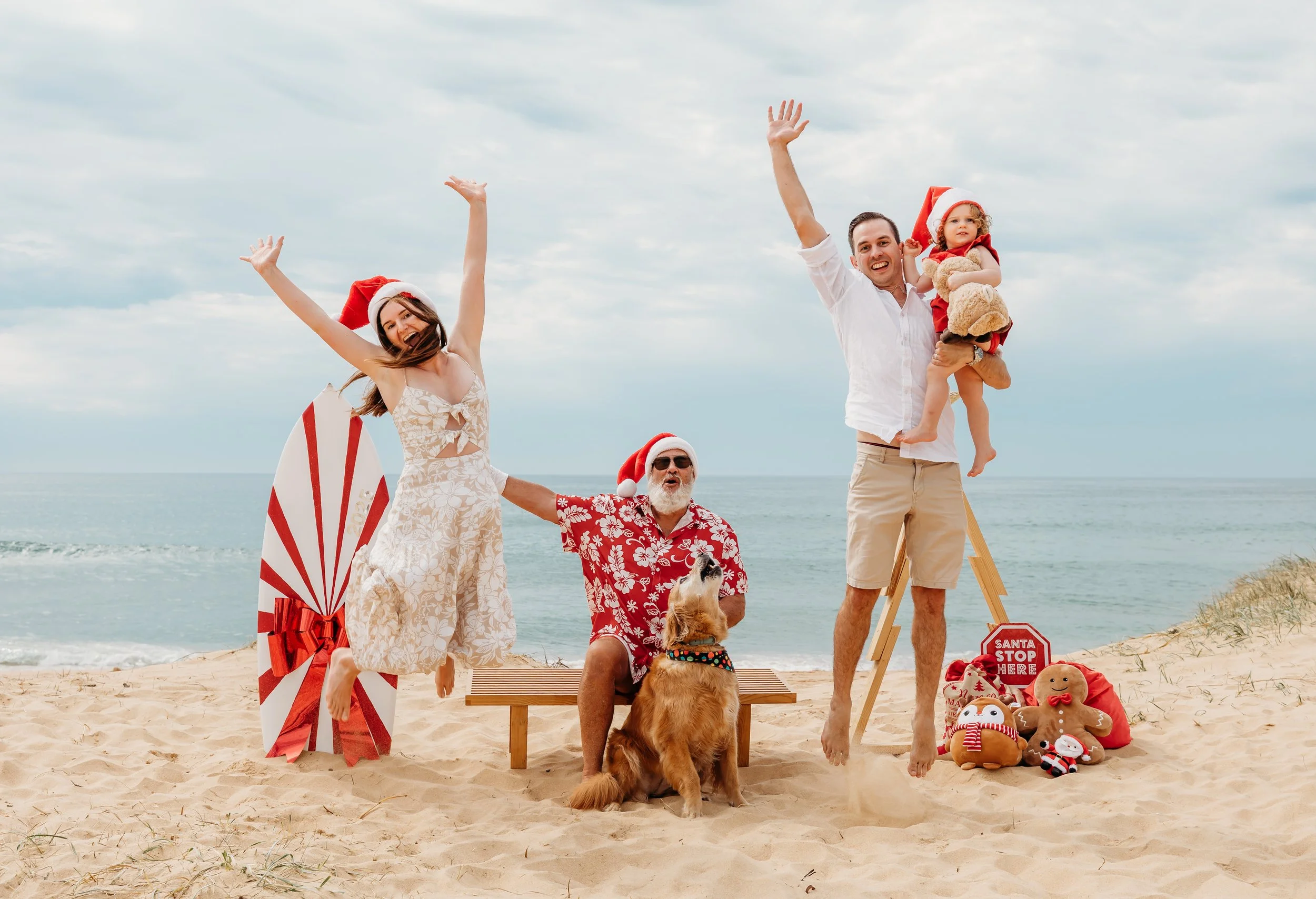 A family celebrating Christmas on the beach with Santa hats, holiday decorations, and gifts, with the ocean and cloudy sky in the background.