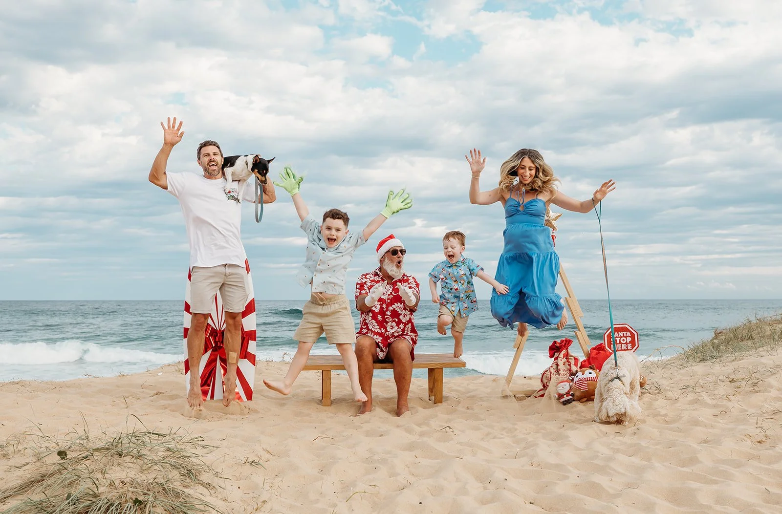 Group of people celebrating Christmas on a beach, including a man with a dog, children, a woman, and Santa Claus, with Christmas decorations and a sign that says 'Santa stop here.'