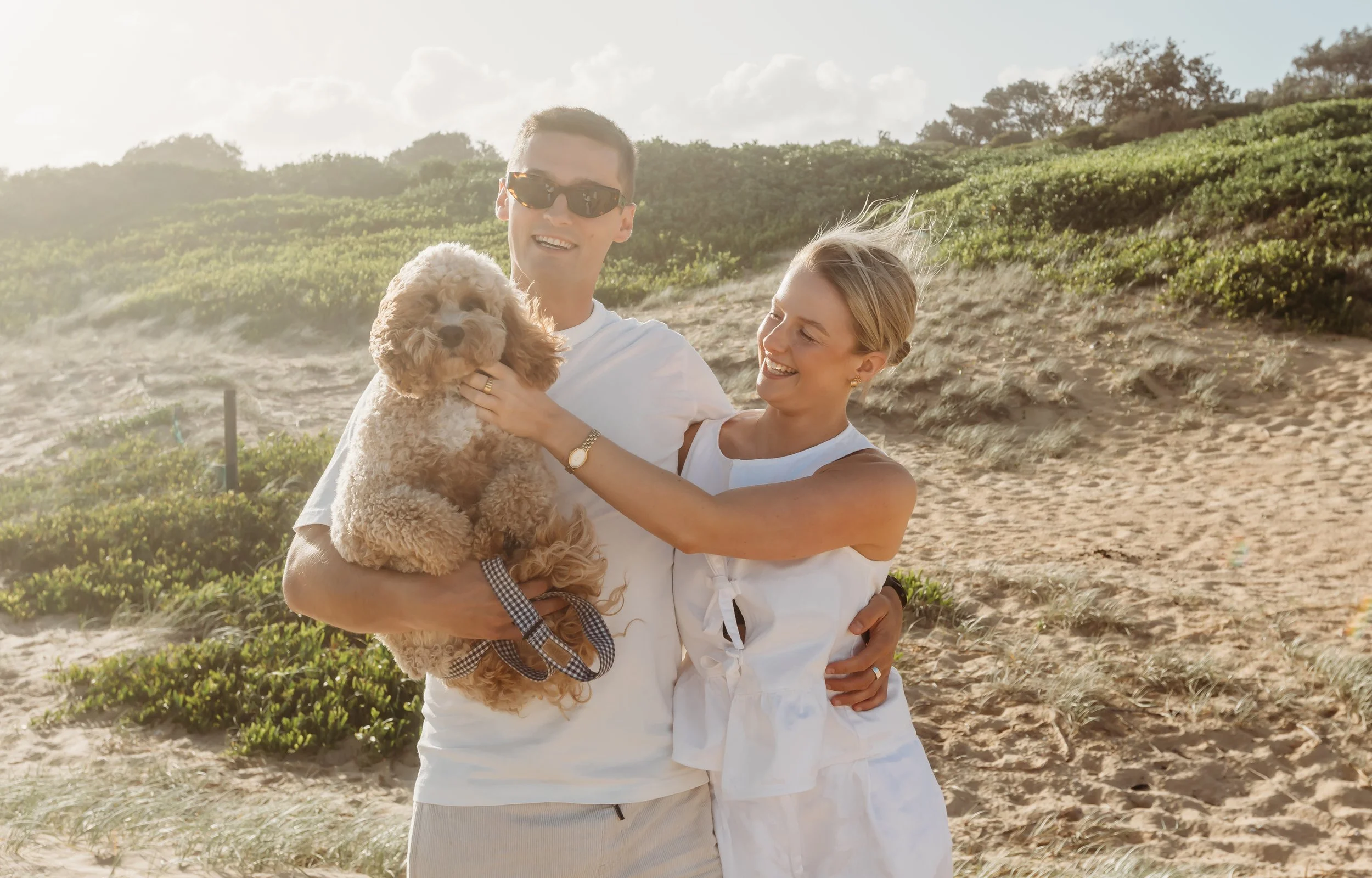 A man and woman smiling at each other on a beach, holding a fluffy dog. The man is wearing sunglasses and the woman has blonde hair. There are sand dunes and green bushes in the background with a partly cloudy sky.