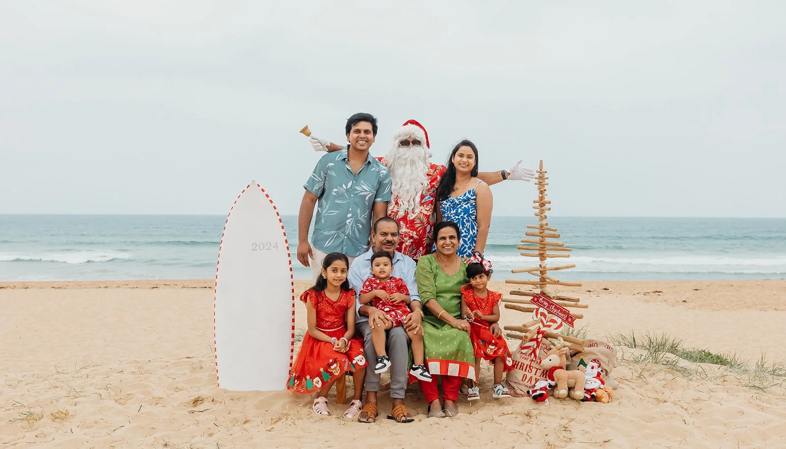 A family dressed in Christmas attire on a beach with Santa Claus, holiday decorations, and a sand Christmas tree, celebrating Christmas day.