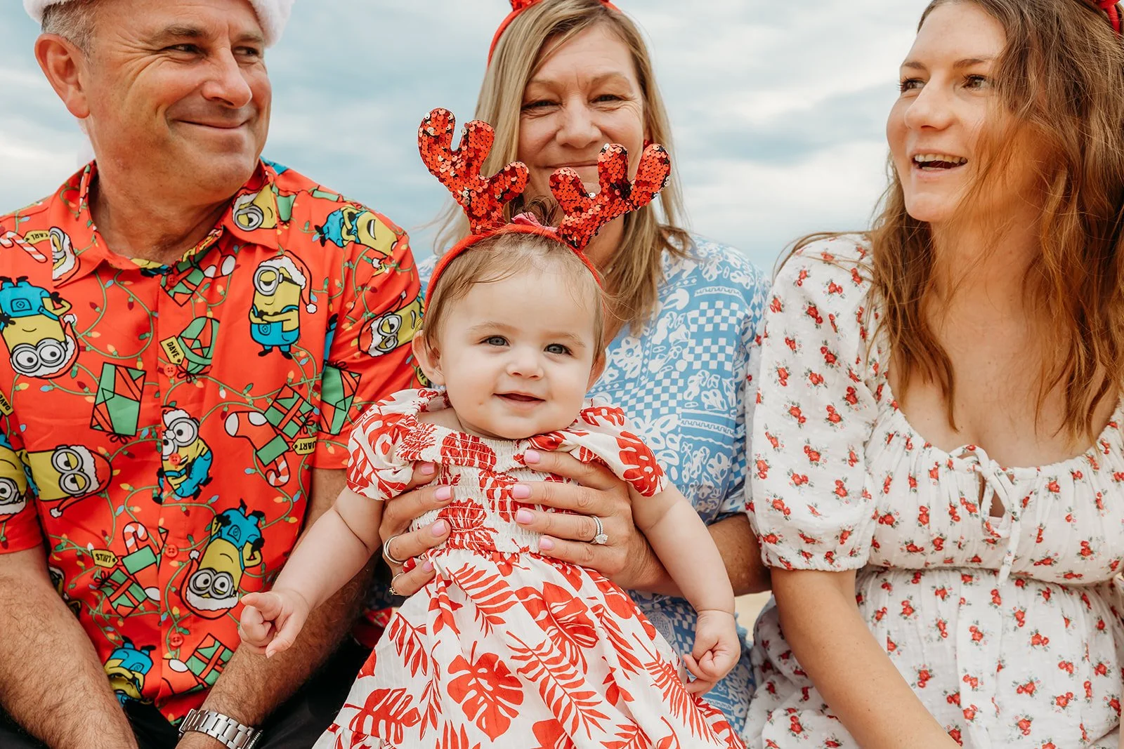 Four people, including a baby girl wearing a red and white dress and red antler headband, are seen close together outdoors. The older woman and man are smiling, while the woman on the right is looking at the woman in the middle. The man is wearing a colorful Disney-themed shirt, and the baby is being held by the older woman.