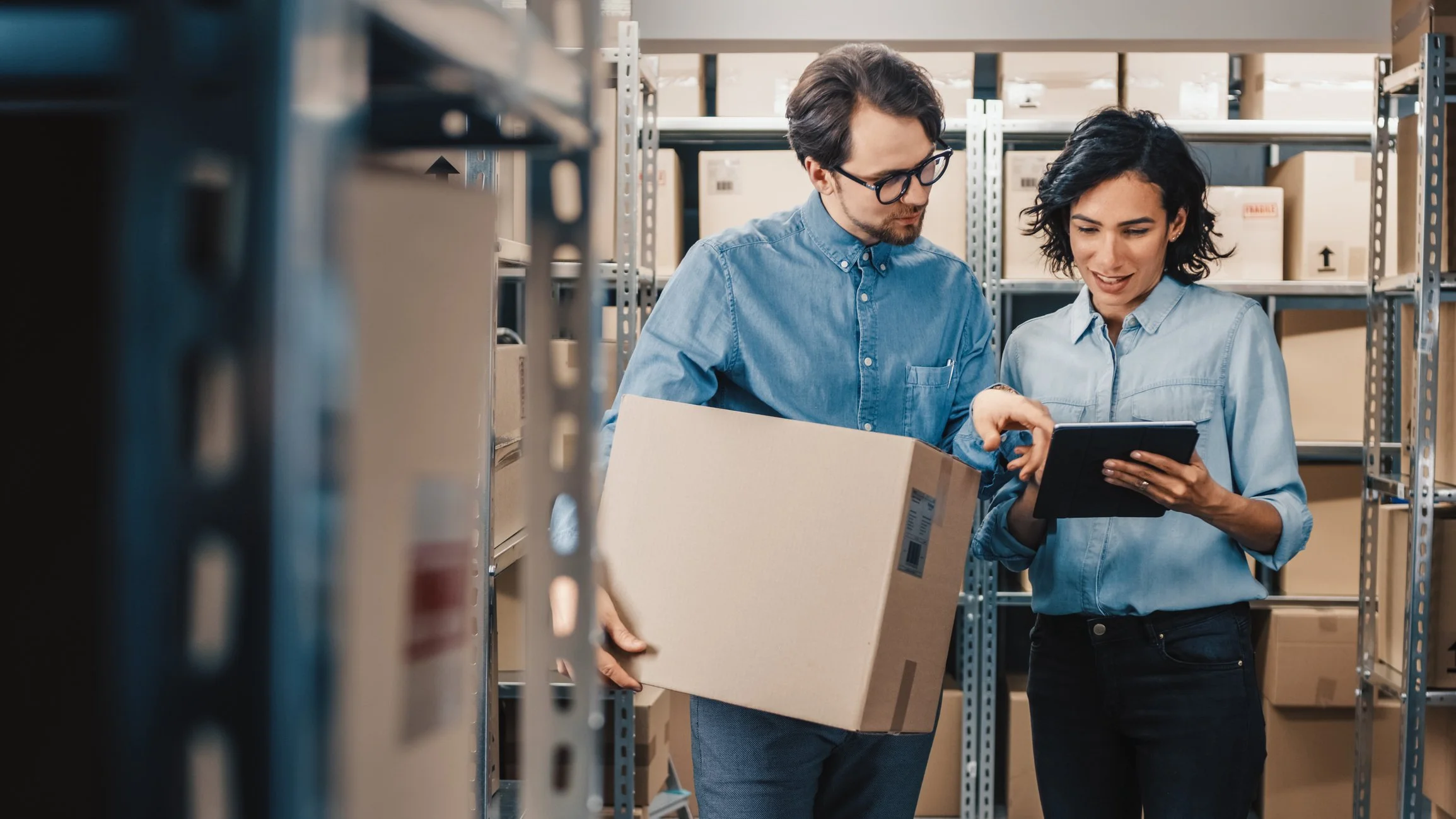 Two people working in a storage room filled with shelves of boxes. The man is holding a box while the woman shows something on a tablet.