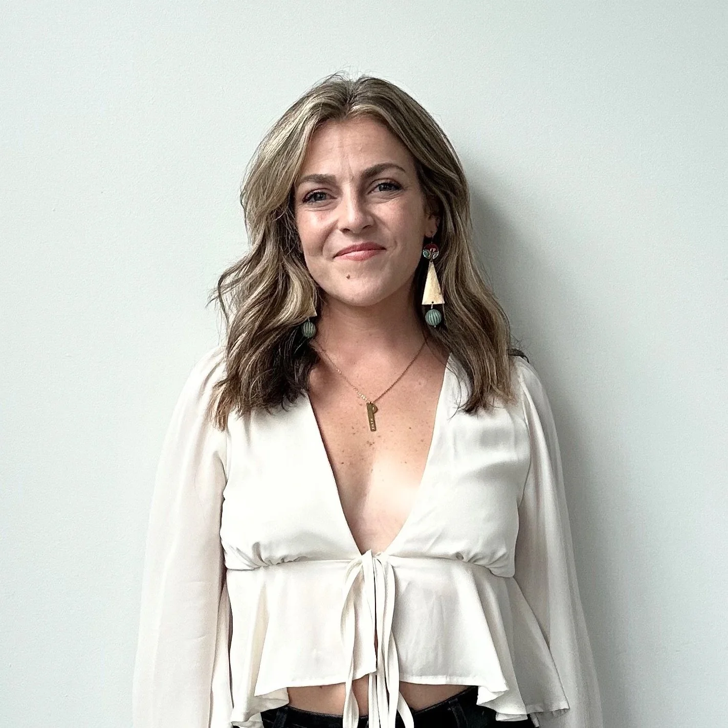 A woman with wavy light brown hair smiling, wearing a white cropped blouse with a tie in front, hoop earrings, and a gold necklace with a pendant, standing against a plain white wall.