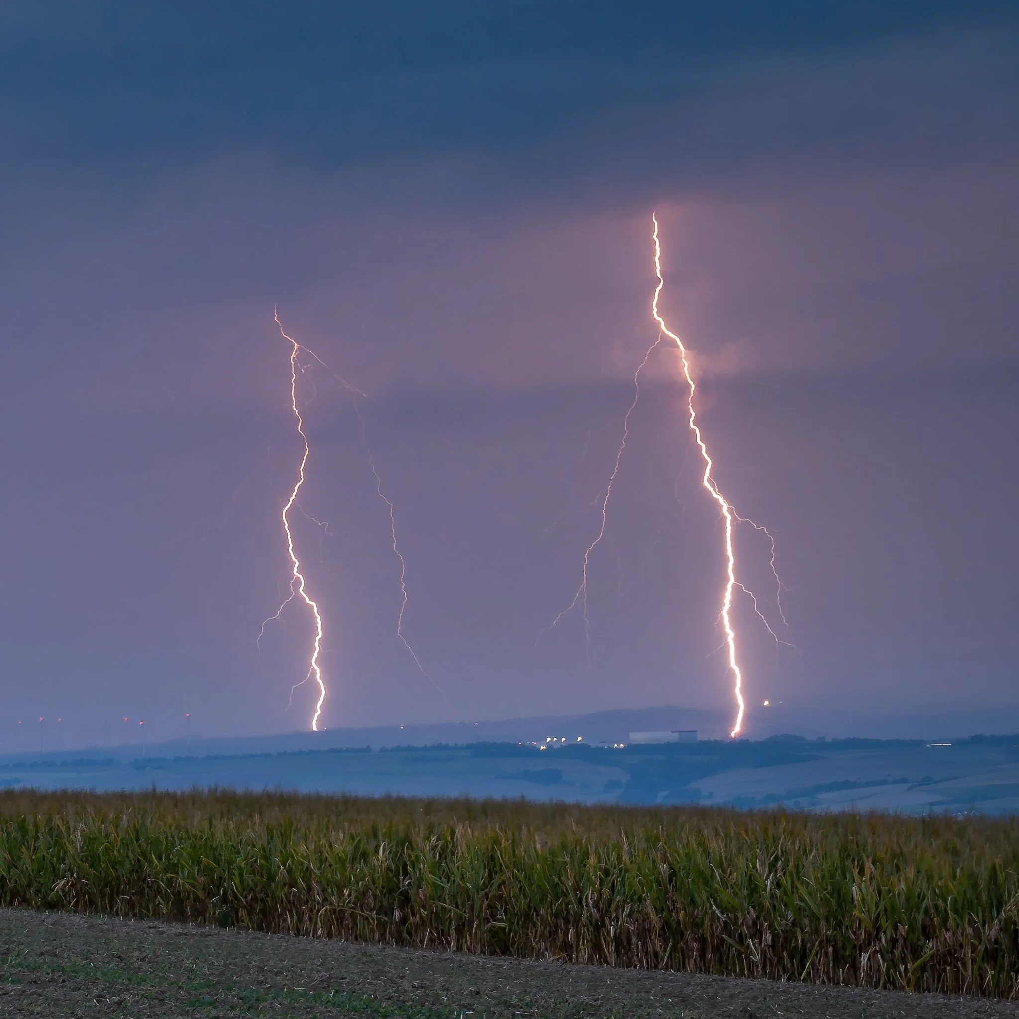 Two large lightning bolts striking down over a rural landscape with fields and hills, under dark stormy sky.