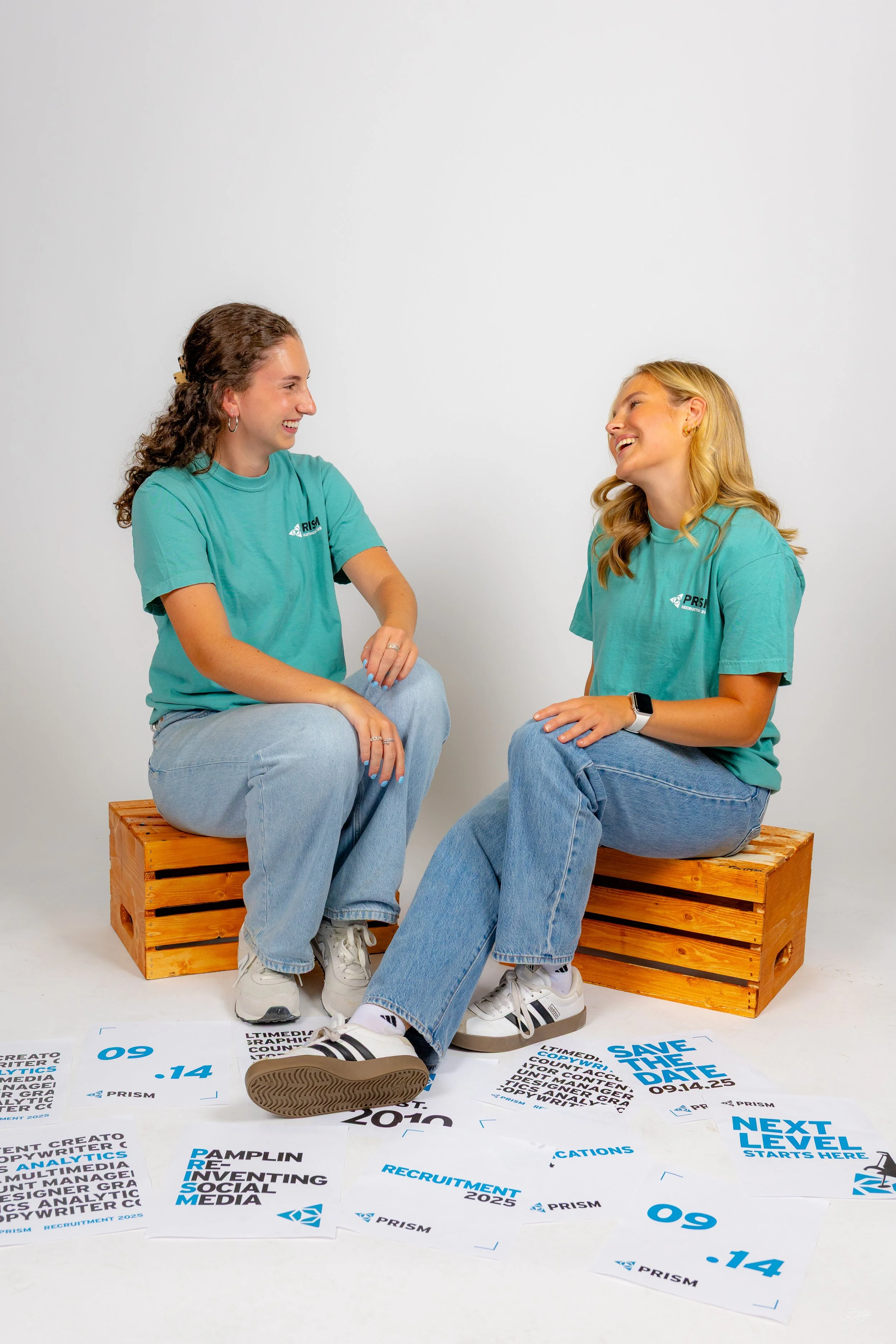 Two young women sitting on wooden crates, smiling and engaging in conversation, wearing teal T-shirts with logos, against a plain white background, with printed papers spread on the floor.