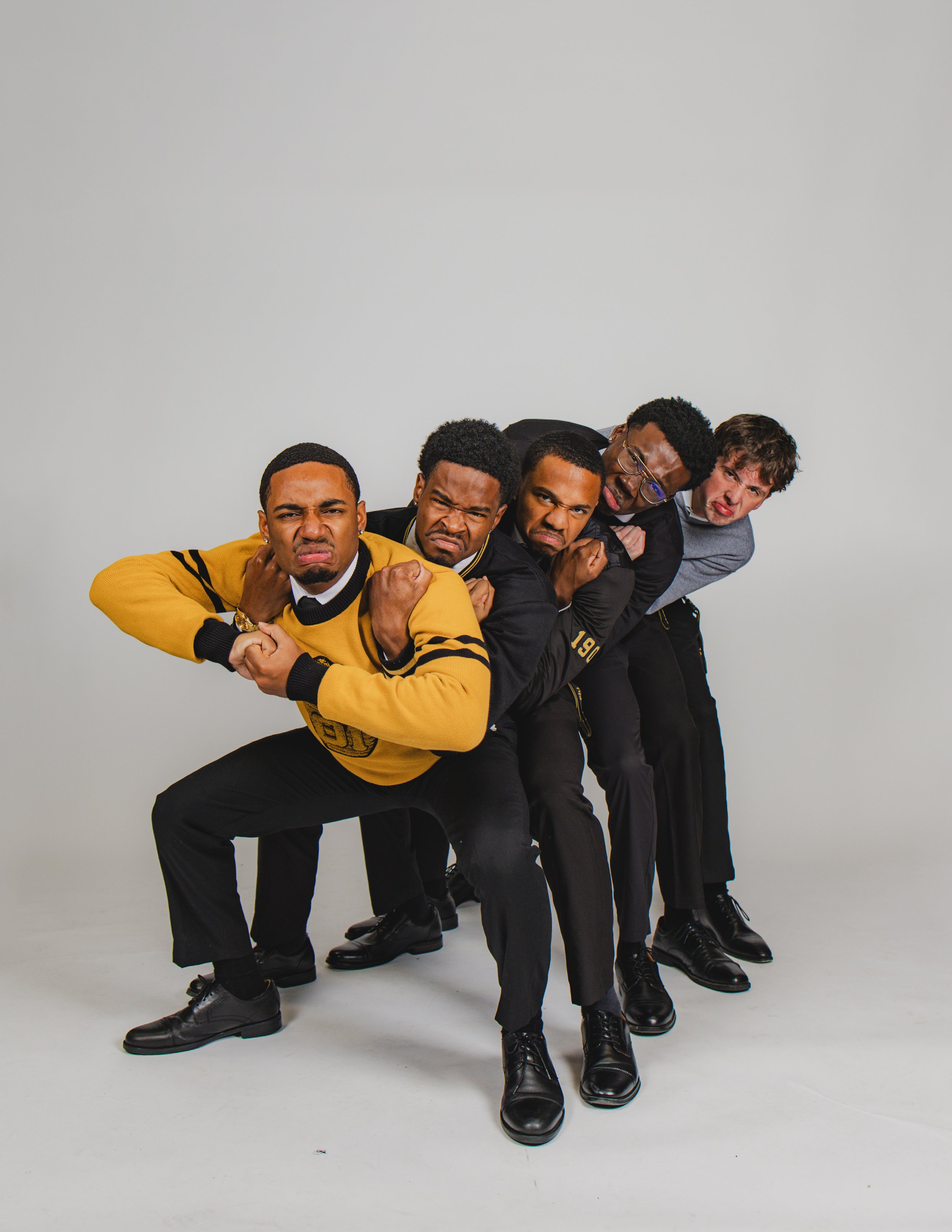 Five young men standing in a line, leaning forward with their arms crossed and looking angry against a plain gray background.