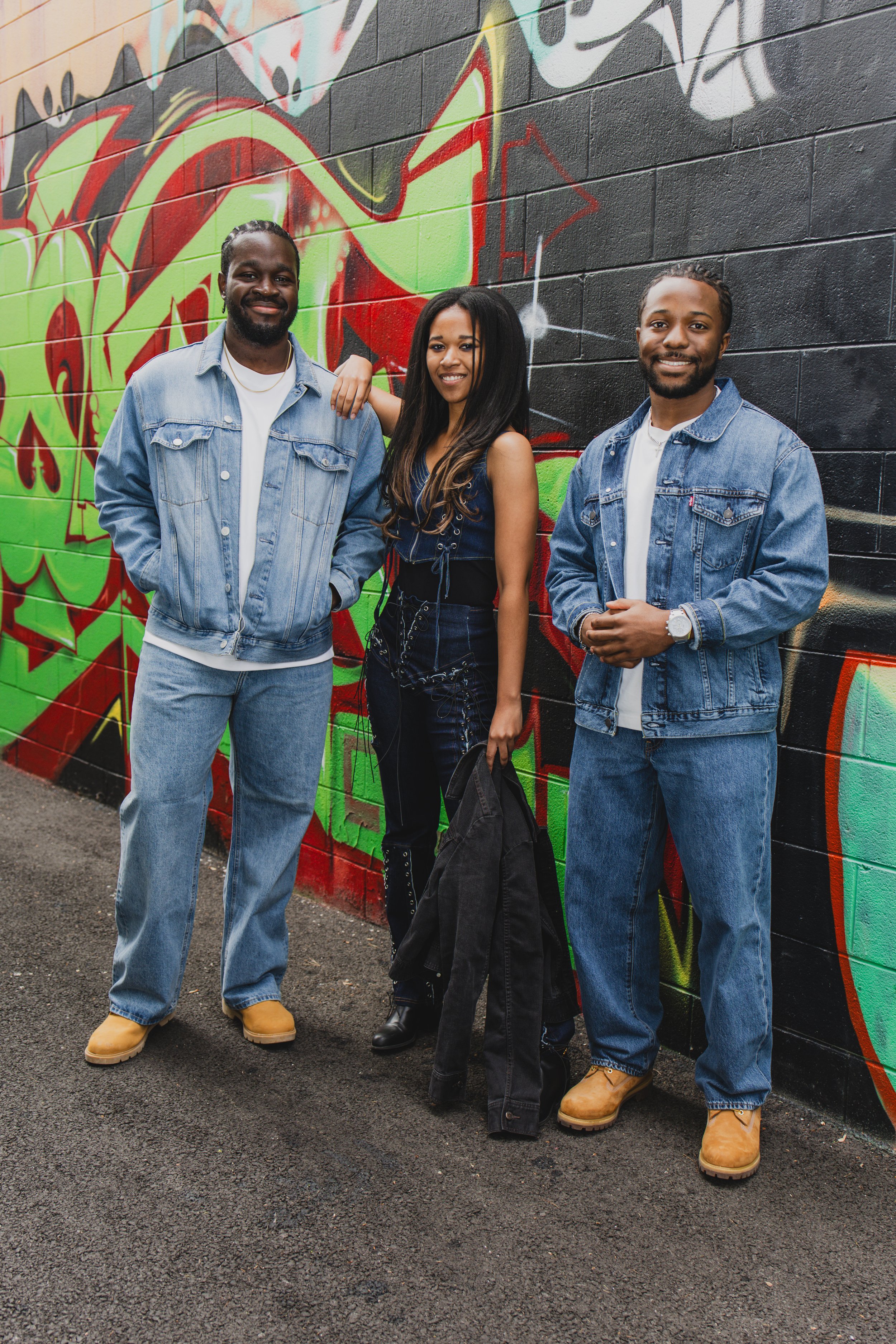 Three young adults standing in front of a colorful graffiti mural on a black brick wall. The person on the left is a man with dark hair and a beard, wearing a denim jacket, light jeans, and tan work boots. The woman in the middle has long dark hair, 