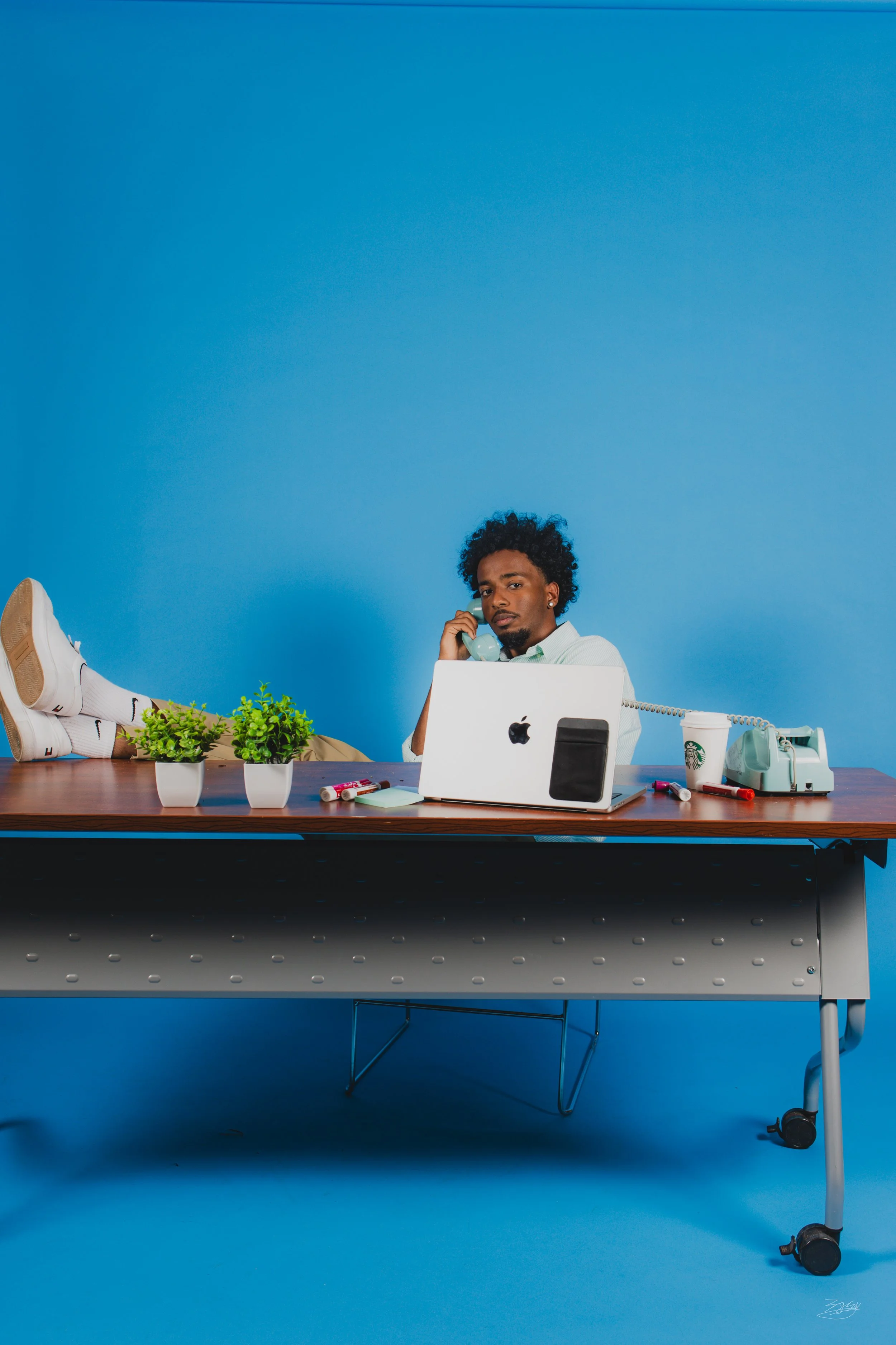 Young man sitting at a desk with plants, a laptop, and vintage phones, against a blue background, with his legs up on the desk, talking on a cellphone.