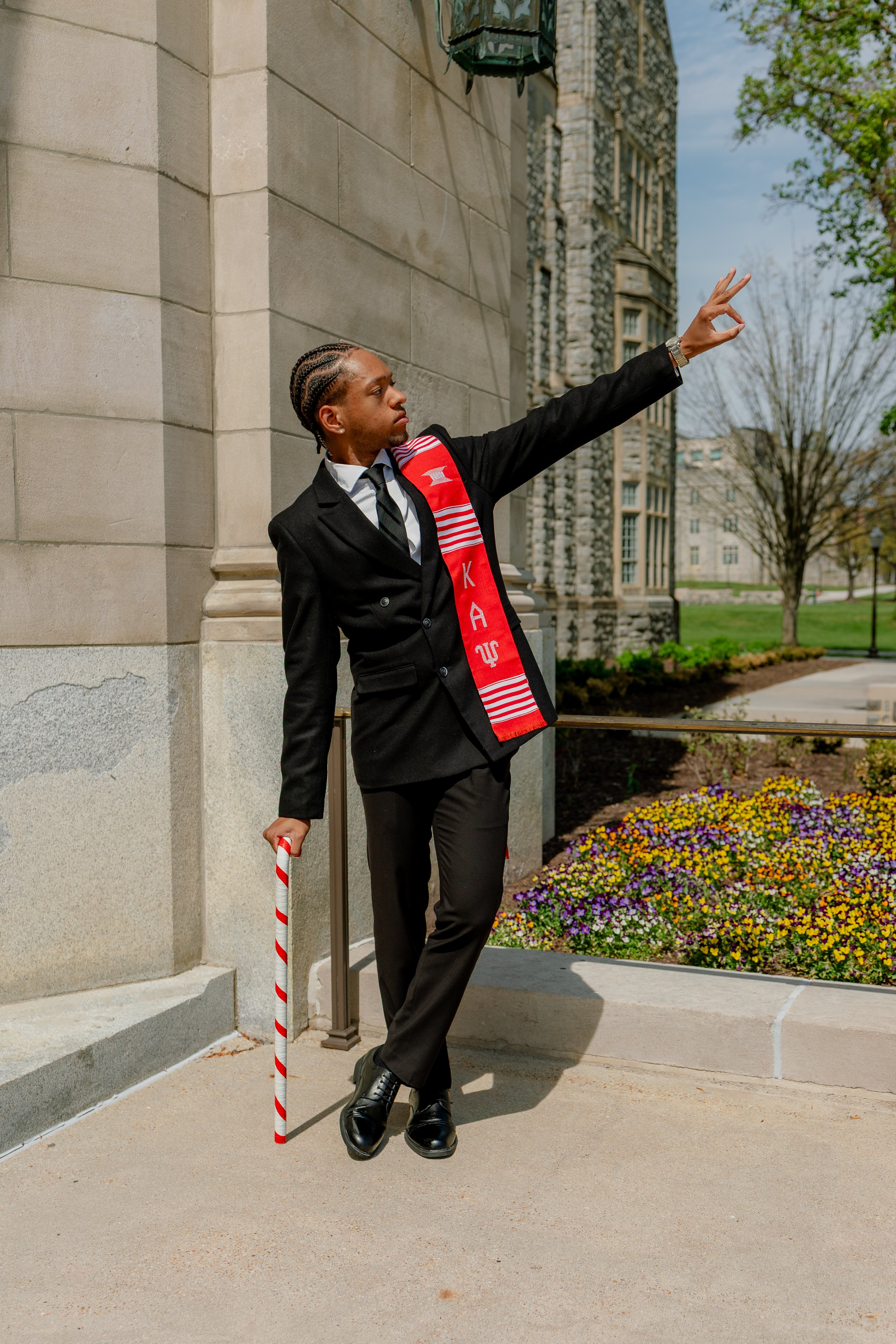 A man dressed in a black suit, white shirt, and black tie stands outside a stone building. He wears a red graduation stole with white and red stripes and the letters 'KAS' visible. He has braided hair, a gold watch, and is holding a candy cane in his
