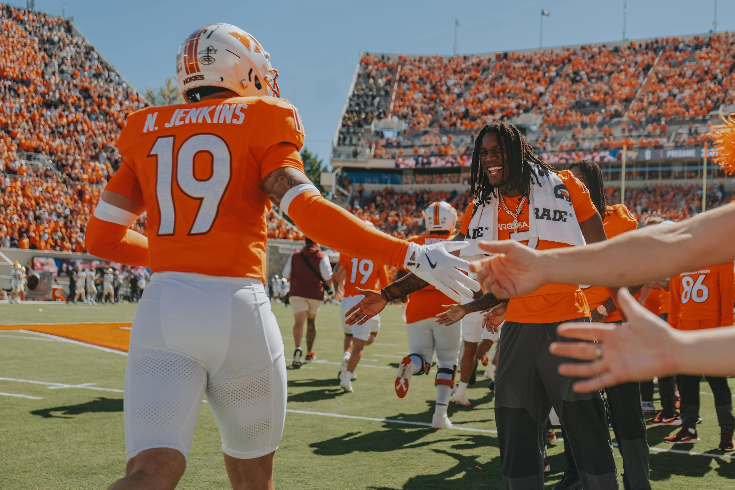 Football players in orange and white uniforms on the field during a game, celebrating and giving high fives, with a crowd in the stands behind them.