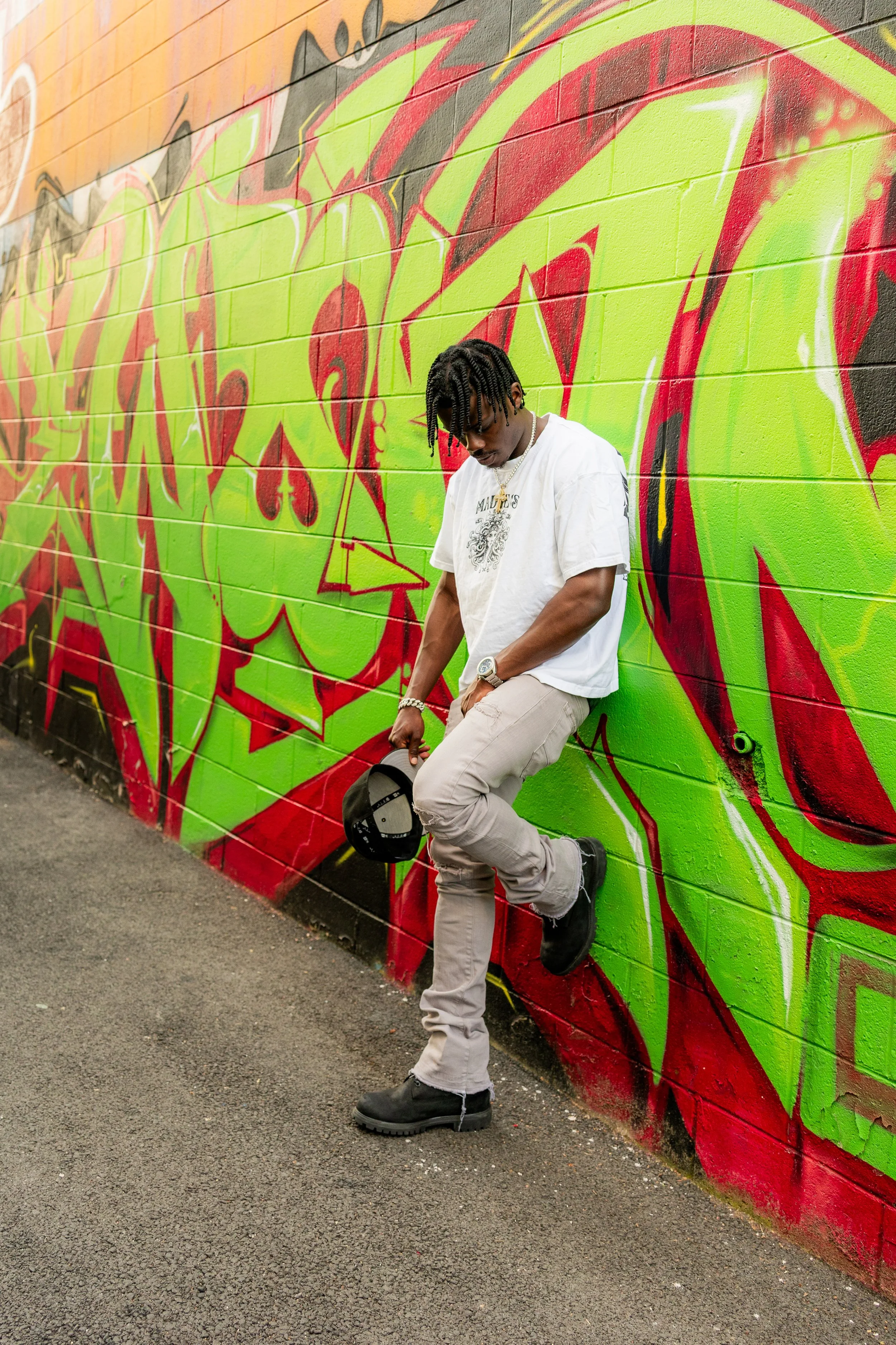 Young man with twisted hair wearing a white t-shirt, beige pants, and black boots, standing against a vibrant graffiti mural on a brick wall. He has a silver watch and a gold chain, holding a hat in his right hand, looking downward.