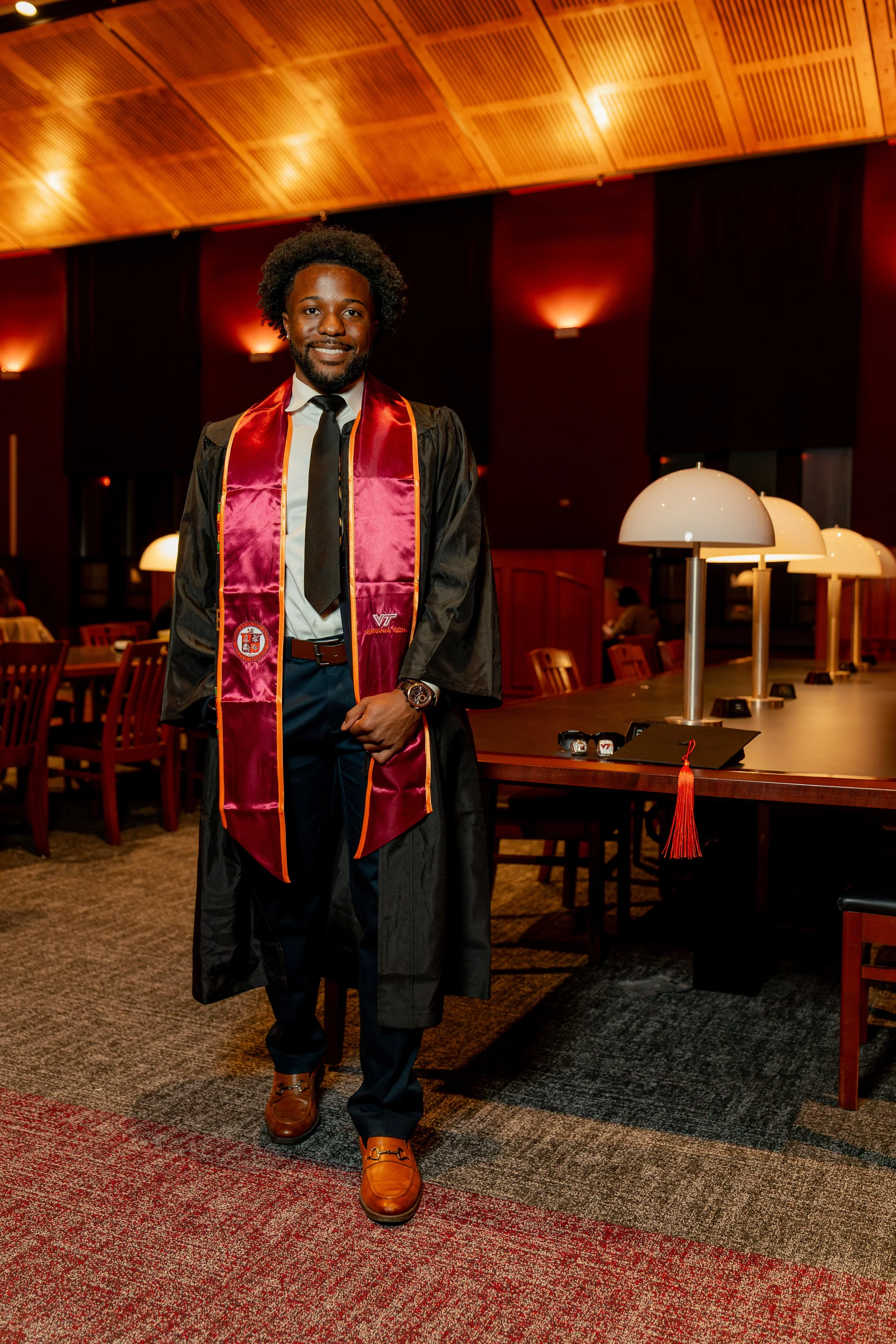 A man in graduation gown and cap smiling at the camera in a decorated indoor setting with tables, chairs, and warm lighting.
