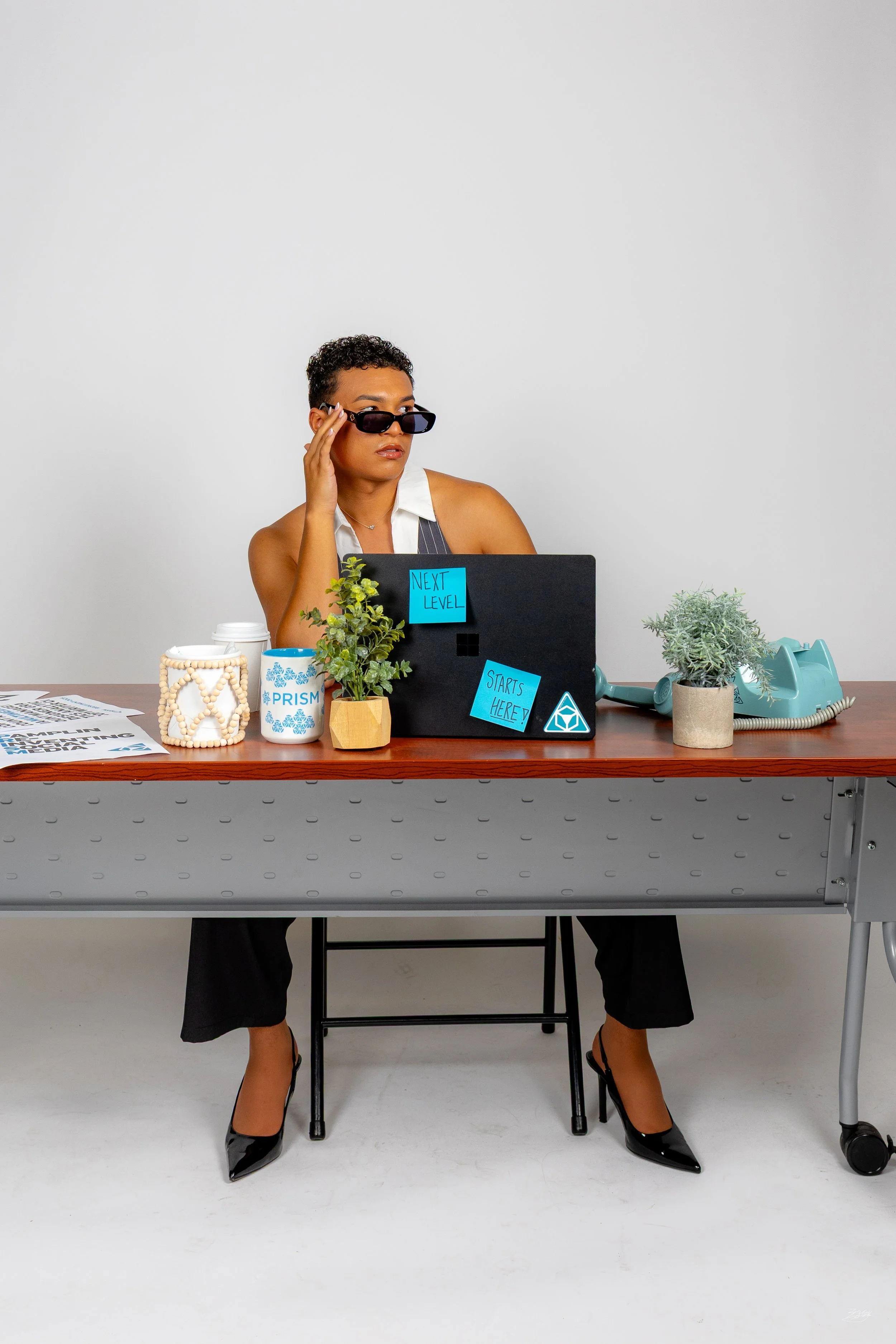 Woman sitting at a desk, wearing sunglasses, black high heels, a white sleeveless blouse, and a pinstripe vest. The desk has plants, a coffee mug, a blue telephone, and various office papers and signs that read "Next Level" and "Starts Here!"