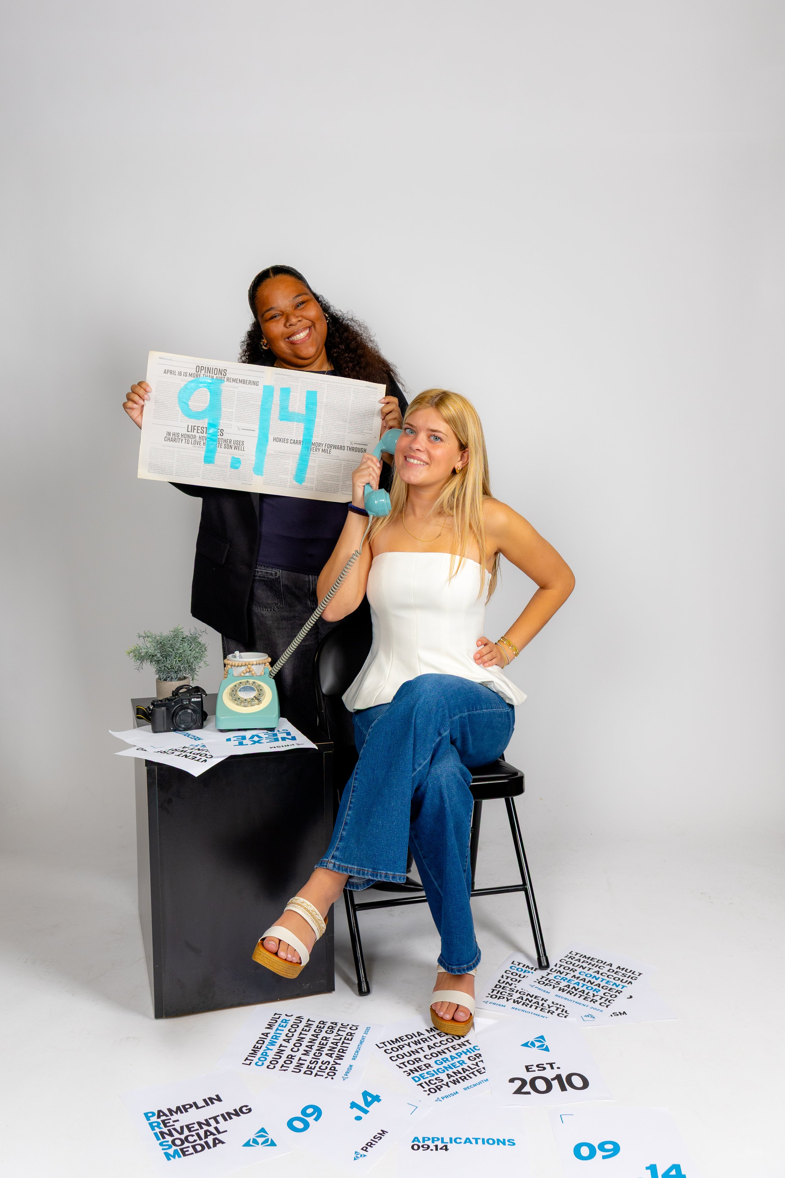 Two women posing in an office studio with scattered posters and promotional materials. One woman is standing, holding a newspaper, and smiling, while the other is sitting on a stool, talking on a vintage rotary phone, with her leg crossed and a smile
