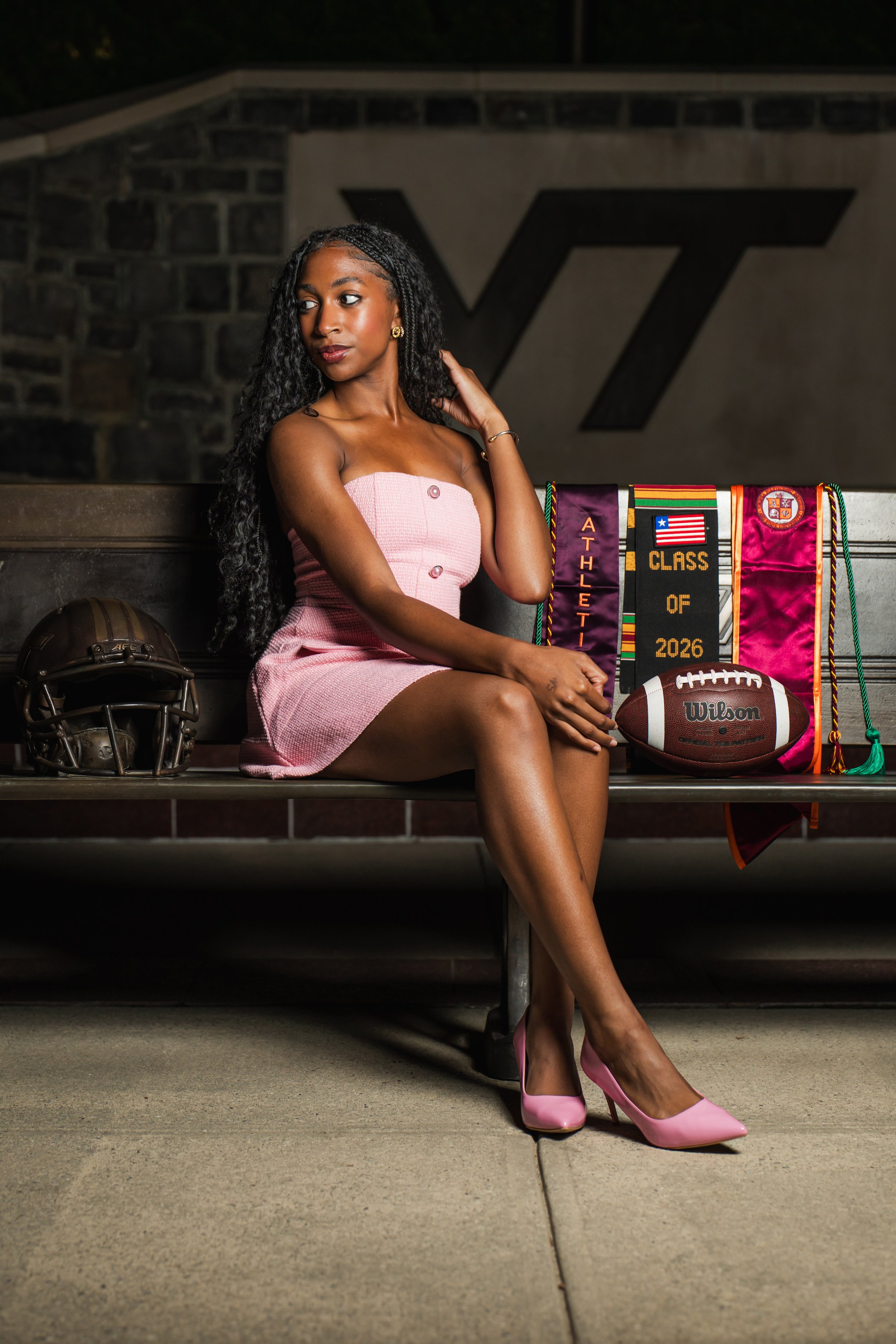 A young woman in a pink strapless dress with button details, pink high heels, and gold jewelry, sitting on a bench at night with football equipment and banners behind her, including a football helmet, a Wilson football, and banners that say 'Athletic