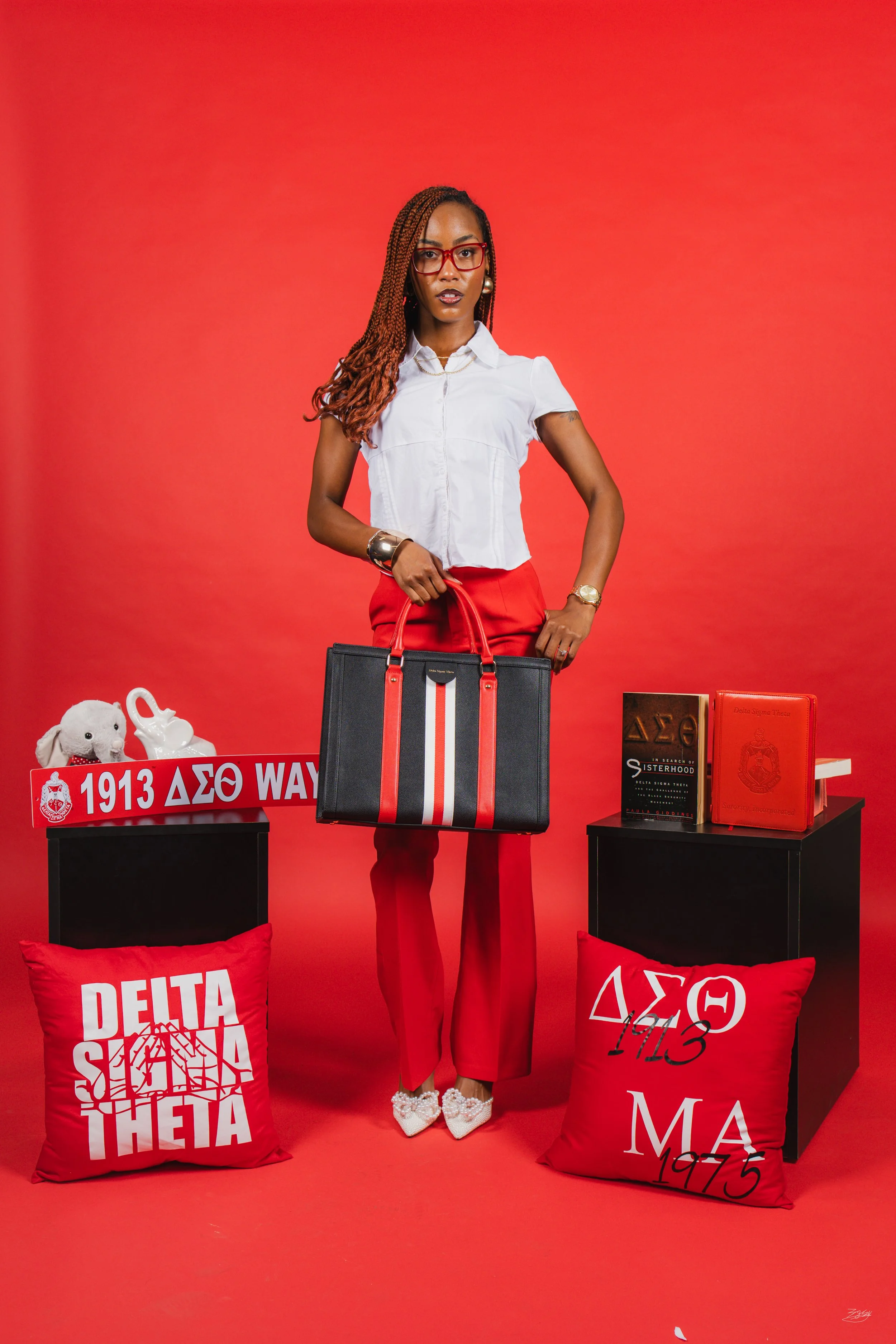 A woman with braided hair, red glasses, and a white shirt standing in front of a red background. She is holding a large black and red tote bag. There are red pillows with white and black Greek letter logos and university crests on black tables, and v