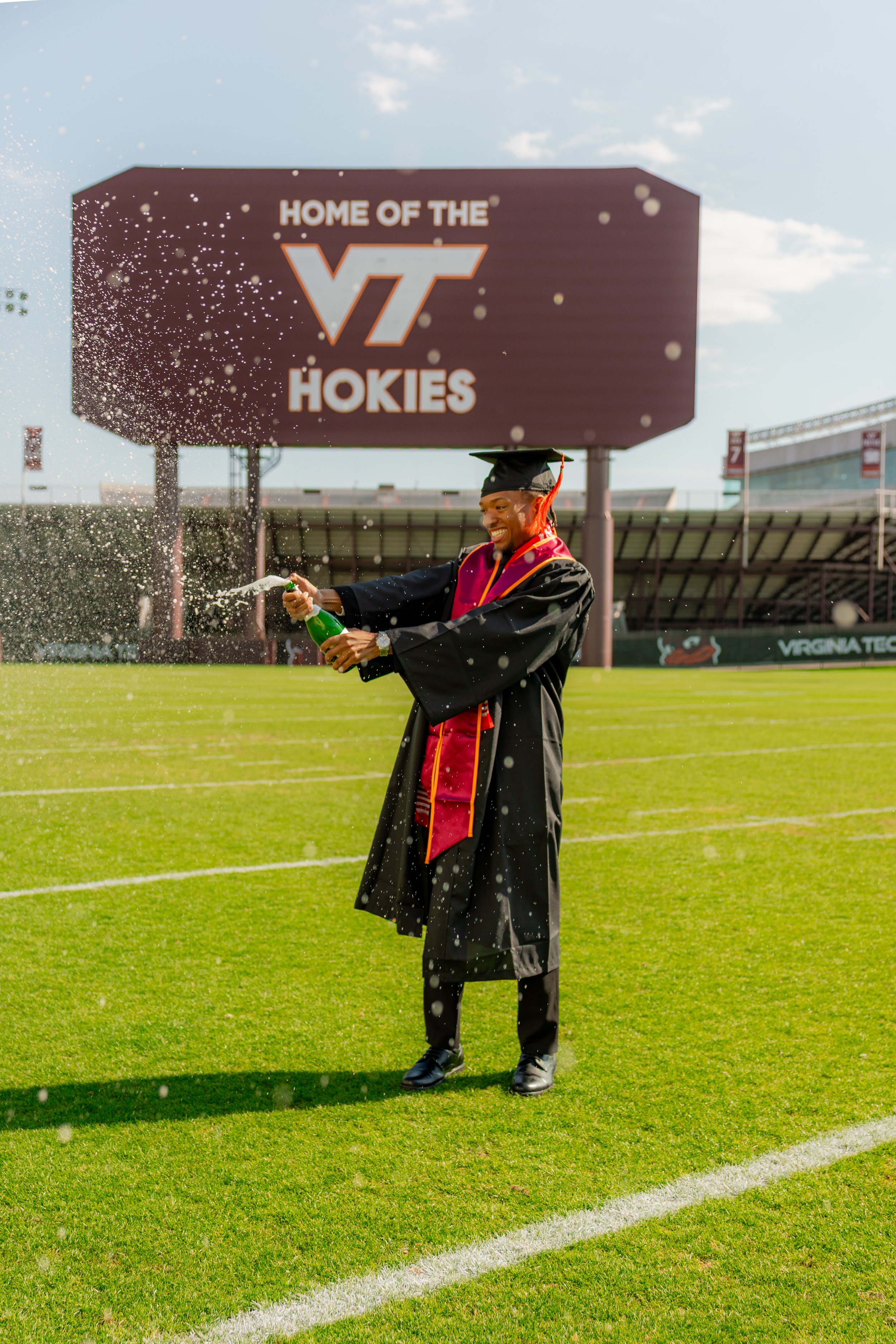 A man in graduation attire spraying champagne on a football field, with a sign in the background that reads 'Home of the VT Hokies'.