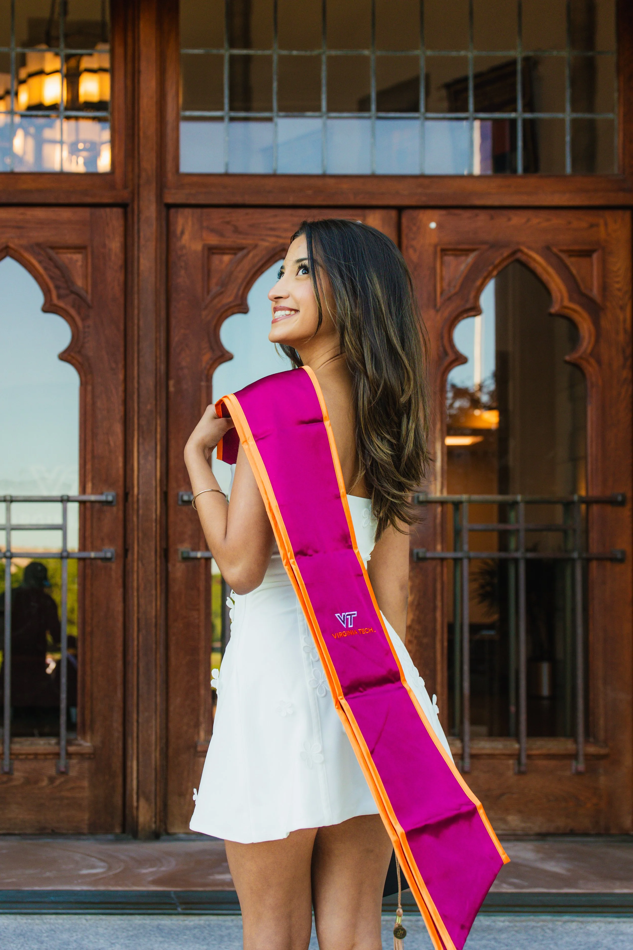 A woman wearing a white dress and a Virginia Tech graduation stole, smiling and looking up, standing in front of a large wooden and glass door.